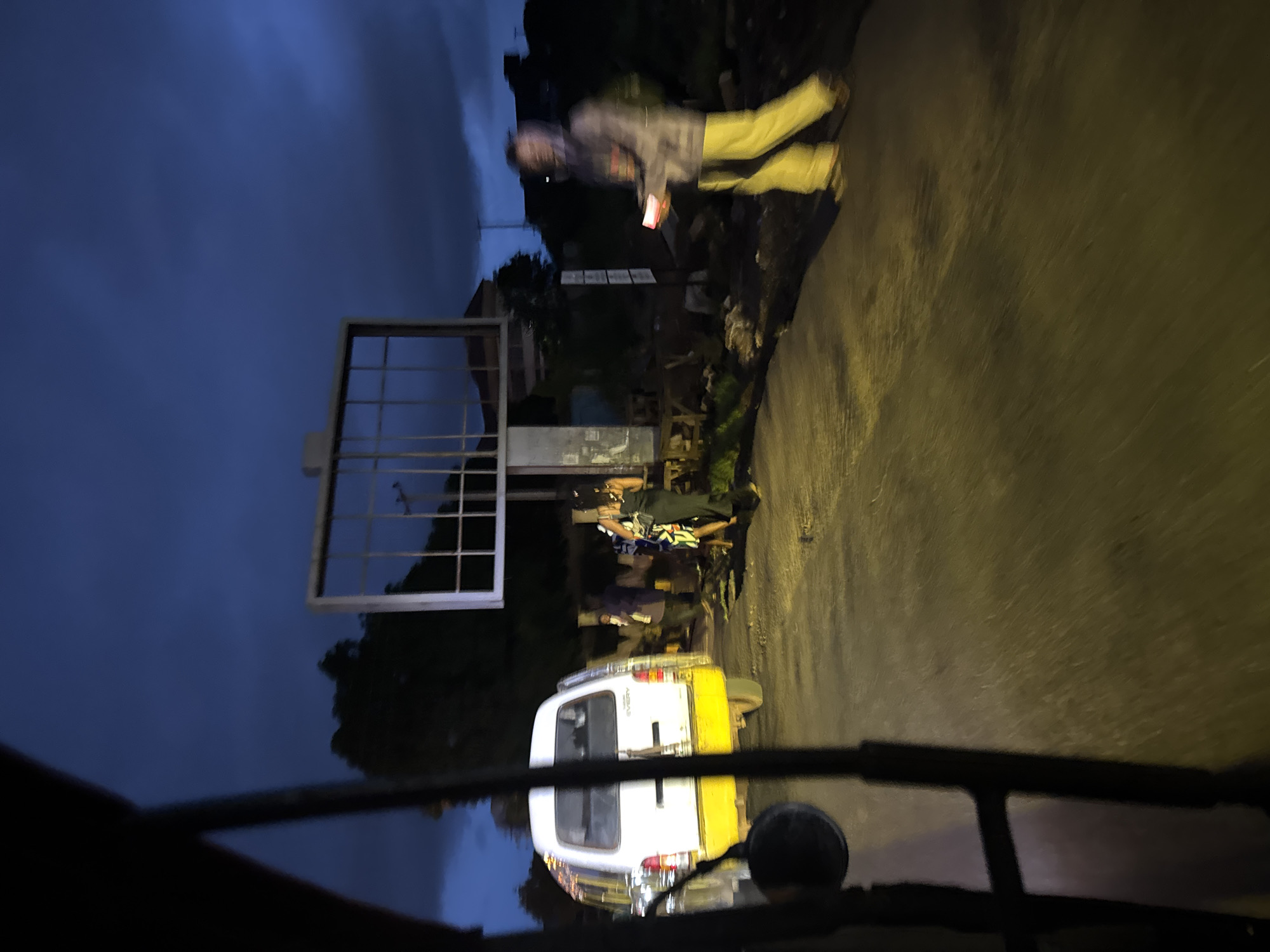 The image depicts a street scene at dusk in Enugu, Nigeria, characterized by a dark, cloudy sky and the presence of street lighting and artificial light sources. In the foreground, a portion of a vehicle, likely a three-wheeled auto-rickshaw or similar transport, is visible on the left, with dark metal bars framing the view.
A white van with a yellow lower body is parked on the right side of the road, facing away from the camera. Several people are gathered near the van. One individual, wearing a grey hooded jacket and yellow pants, is walking from right to left across the road, holding a glowing rectangular object, possibly a phone or a small light. Behind the van, a group of people are standing and interacting. One person in the group is wearing a black dress, and another is in a blue and white patterned outfit.
Further in the background, a large, empty billboard structure stands prominently, with a grid-like frame. The surrounding environment shows signs of urban infrastructure, with some vegetation and potentially other buildings or structures obscured by the low light conditions. The road surface appears to be paved and wet, reflecting the available light. The overall impression is a typical evening scene in a Nigerian city, with a sense of movement and activity despite the fading light.