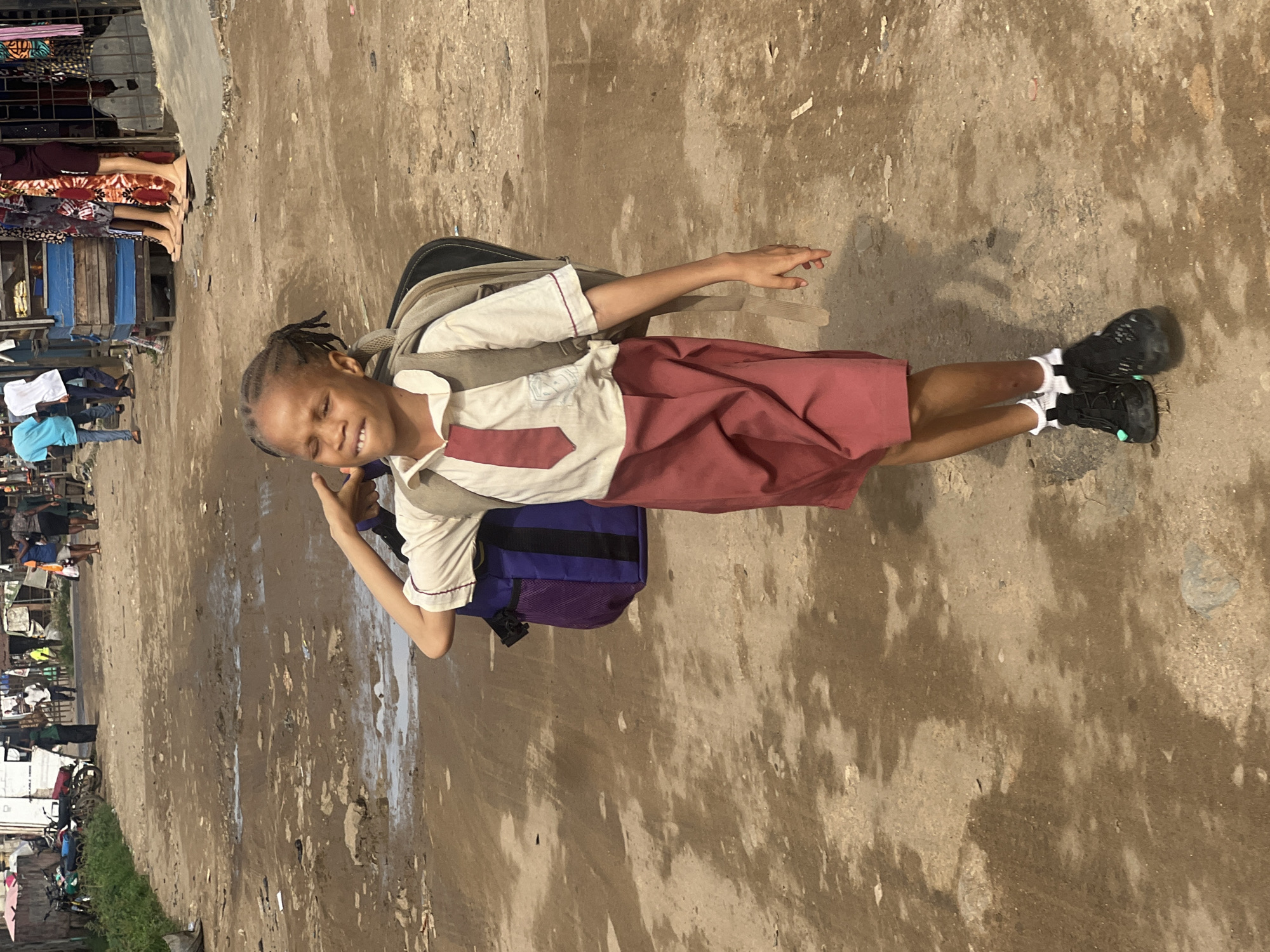 A young girl, likely a student, is the central figure in this daytime outdoor shot taken on a street in Ijegun, Nigeria. She stands on a rough, unpaved or poorly paved road that appears muddy or wet in patches, with varied shades of brown, tan, and grey.
The girl has dark skin and her hair is styled in braids. She wears a school uniform comprising a light-colored collared shirt with maroon accents and a tie-like embellishment, and a maroon knee-length skirt. A large backpack, a combination of tan, blue, and purple, is strapped to her back. She completes her attire with white socks and black athletic shoes that feature a subtle light green detail near the toes. Her face is turned towards the camera, beaming with a wide, joyful smile, her eyes squinted, possibly due to bright sunlight.
In the background, to the left of the girl, is a bustling street scene with several market stalls or temporary shops displaying various fabrics and goods. People are visible near these stalls, engaged in daily activities; one person wears a light blue shirt, another a white top, and others are partially obscured. Further down the road, several motorcycles or scooters can be seen parked. A narrow strip of green grass lines the far left edge of the scene.
The bright lighting suggests it is daytime. The girl's cheerful expression and uniform indicate she is either on her way to or from school, bringing a sense of youthful energy to the busy market environment. There is no visible text in the image.