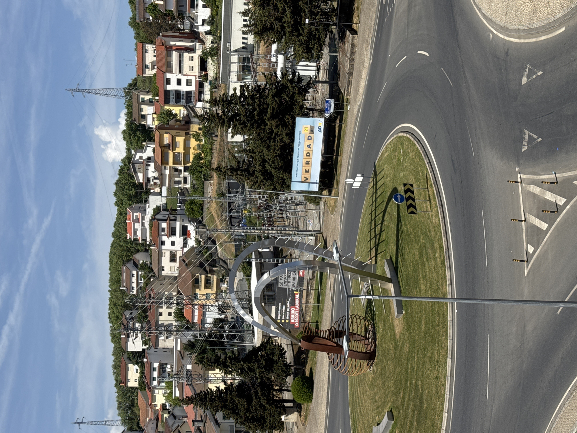 This is an outdoor, eye-level photograph depicting a cityscape with a traffic roundabout in Bragança, Portugal.
The roundabout has a grass-covered center island with road signs. A modern art installation made of metal curves is located near the left side of the island. A billboard with the word "VERDADE" (meaning "truth" in Portuguese) is visible on the side of the road. A second sign is partly visible, displaying election slogans and other political messaging.
In the background, there are residential buildings of various colors and architectural styles, interspersed with trees. Electrical pylons and power lines can be seen crisscrossing the sky and the landscape between the buildings. The sky is blue with sparse clouds. The photo is taken during daylight, with good natural lighting.