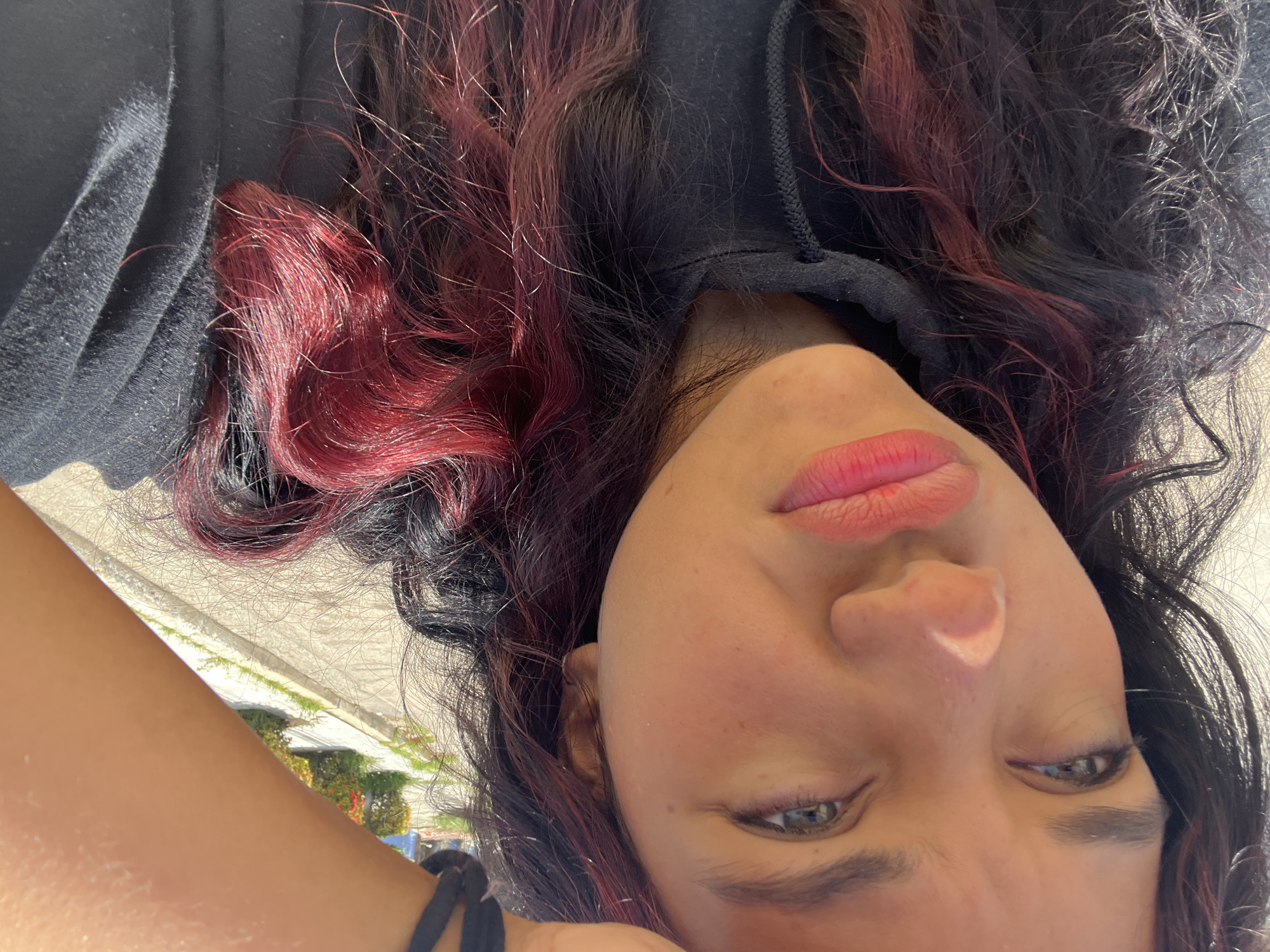 A young person with dark hair streaked with red is lying down, with their face angled towards the camera. They are wearing a black hoodie. The person's eyes are open, and they appear to be looking directly at the viewer, with a neutral or slightly contemplative expression. The lighting suggests it is daytime, possibly late afternoon or early morning, given the strong shadows. The background is blurred but appears to be an outdoor setting, with hints of pavement, greenery, and possibly buildings, consistent with a location in Pampatar, Venezuela. There is no visible text in the image. The overall impression is that of a casual, informal portrait.