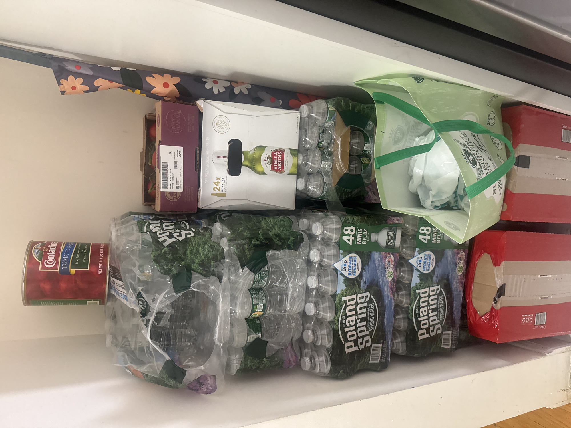 This media file shows a shelf filled with various grocery items, likely in a home pantry or storage area. The setting appears to be indoors, with a white shelf and a dark surface above. The items are arranged in stacks and rows.
On the left side of the shelf, a can of Contadina Tomato Paste is prominently displayed. To its right, a cardboard box containing Stella Artois beer is visible, with "24x BOTTLES" and "5.0% ALC/VOL" printed on it. Inside the beer box, several bottles are packed in plastic.
In the center of the shelf, multiple packs of Poland Spring bottled water are stacked. One pack is labeled "48 MINIS 8 FL OZ BOTTLES" and features an image of a natural landscape. Another pack of Poland Spring water is also visible, with the text "100% NATURAL SPRING WATER".
To the right, a green reusable shopping bag is open and filled with white plastic bags, possibly from a grocery store. Next to it, a red cardboard box with the Coca-Cola logo is present, indicating another beverage item. The overall scene suggests a well-stocked pantry with a variety of beverages and food items. The lighting indicates it is daytime.
Visible text includes brand names like "Contadina", "Stella Artois", "Poland Spring", and "Coca-Cola". Nutritional and product information such as "TOMATO PASTE", "SPRING WATER", "MINIS", "BOTTLES", and "ALC/VOL" are also legible. A date stamp "JUN 16" and other production codes are also visible on some packaging.