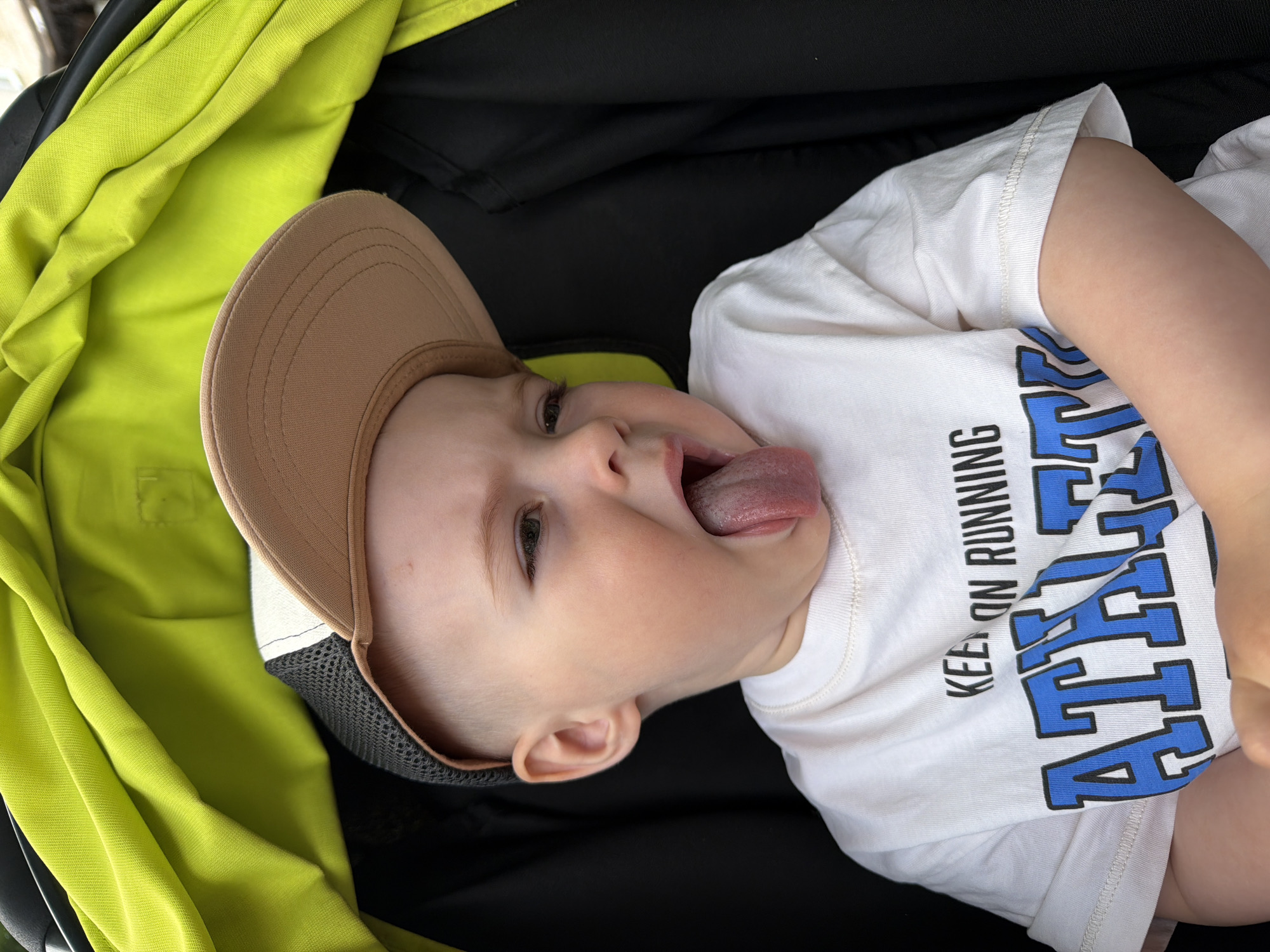 A young child is sitting in a stroller, wearing a white t-shirt and a baseball cap. The child is sticking their tongue out and looking to the right. The stroller has a bright green canopy. The background is dark and out of focus. No specific location in Votkinsk, Russia can be determined from the image.