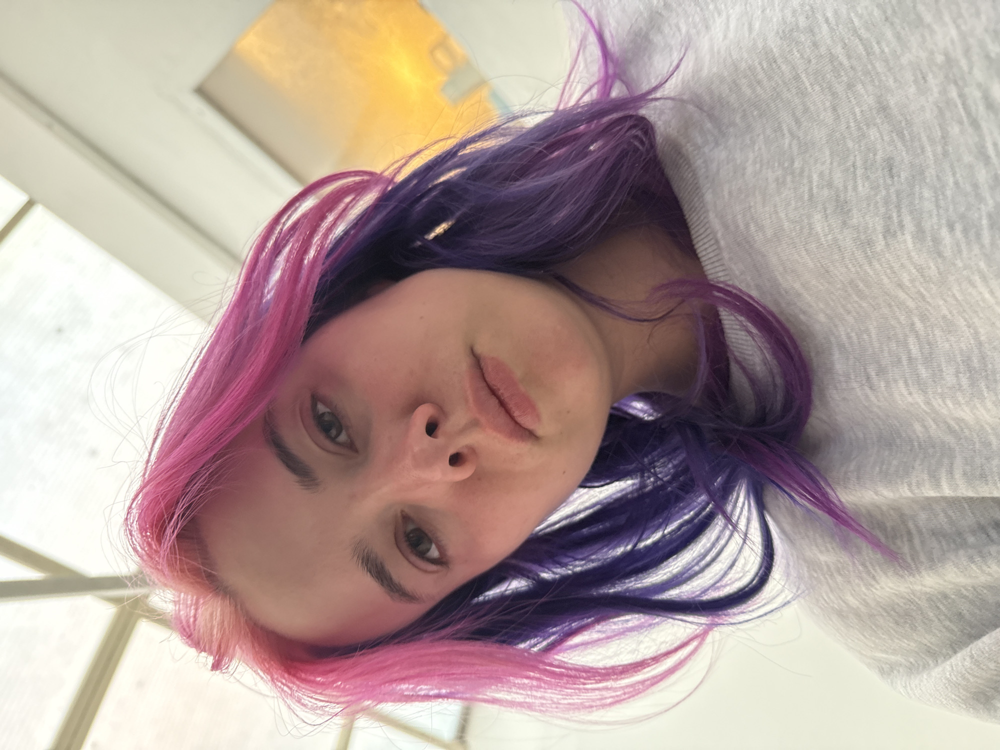 A young woman with pink and purple hair is looking down at the camera. She is wearing a light grey sweatshirt. The background shows a window with sunlight streaming in. The location cannot be determined based on the image.