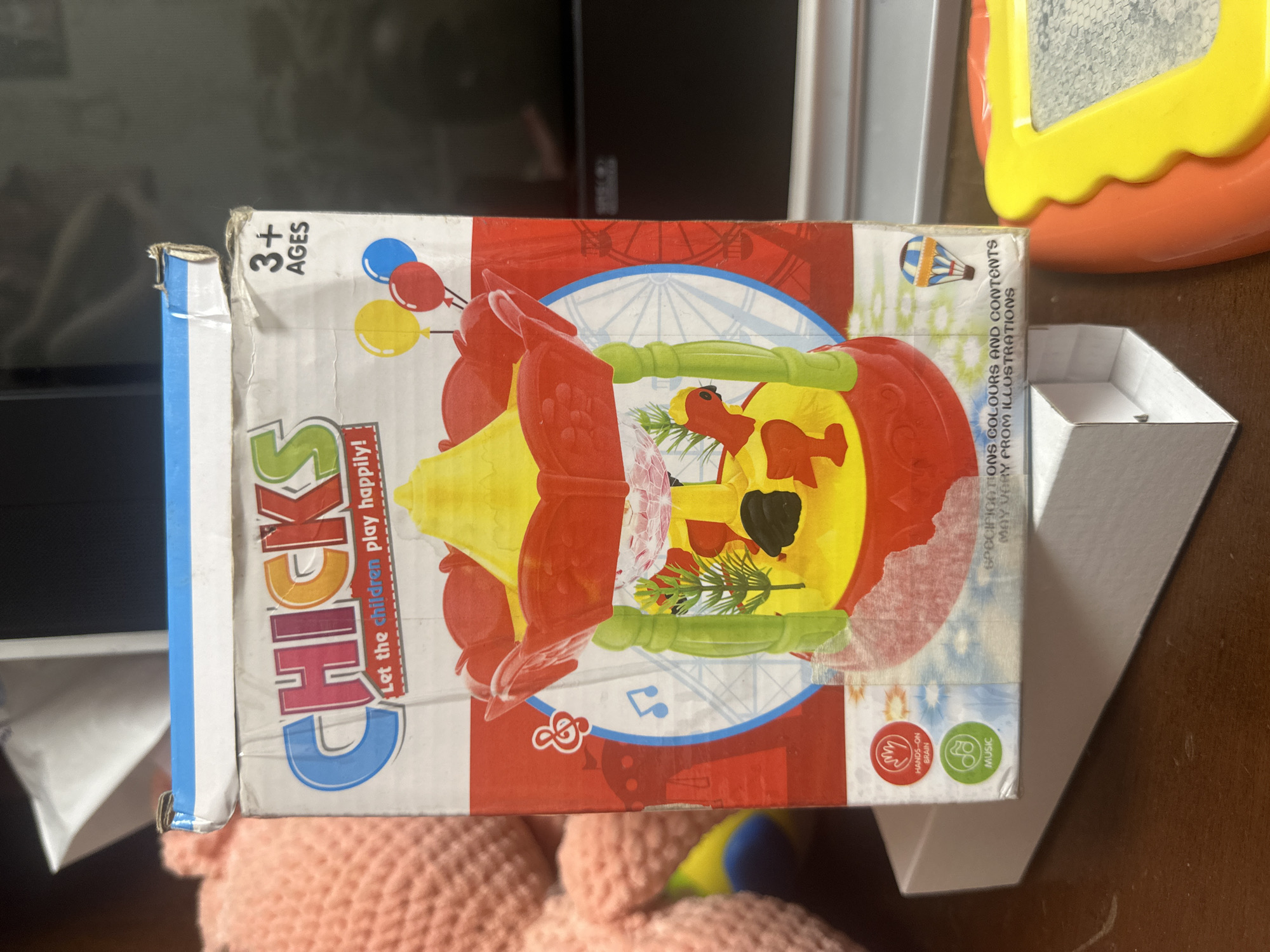 The image shows a toy box for a product called "CHICKS" with the tagline "Let the children play happily!". The box is red and yellow with illustrations of chicks, balloons, and a carousel. The text on the box indicates it is for ages 3 and up and features "Hands-on Brain" and "Music" icons. The box is positioned on a wooden surface, possibly a desk or table. In the background, a blurred pink stuffed animal and a black electronic device are visible. There is no indication of the location being in Загородный, Russia, or any other specific city.