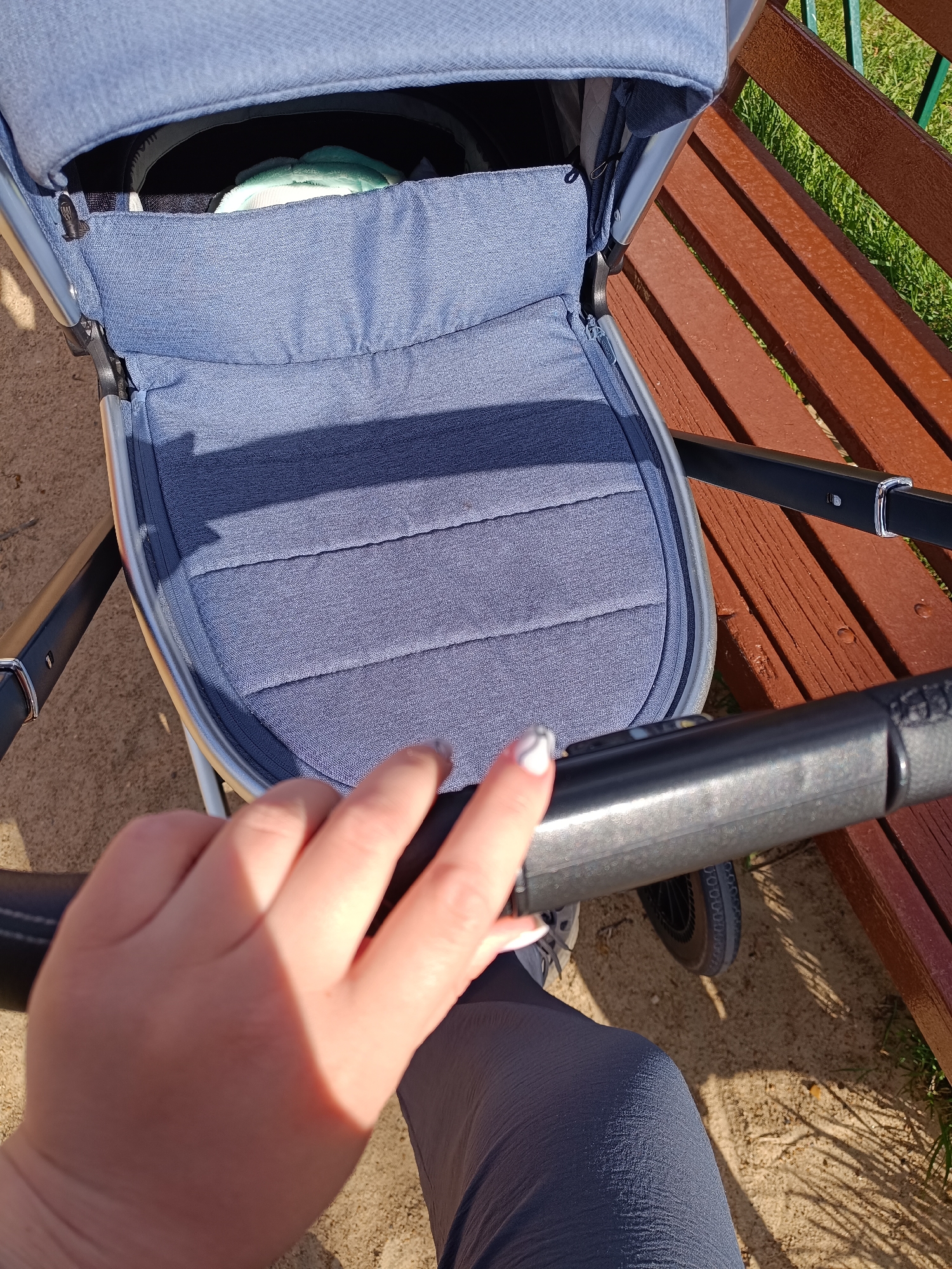 A person's hand rests on the handlebar of a blue baby stroller. The stroller's seat is visible, containing a soft object inside. The stroller is positioned next to a wooden bench. The ground appears to be a mixture of sand and grass. The location might be Tobolsk, Russia.