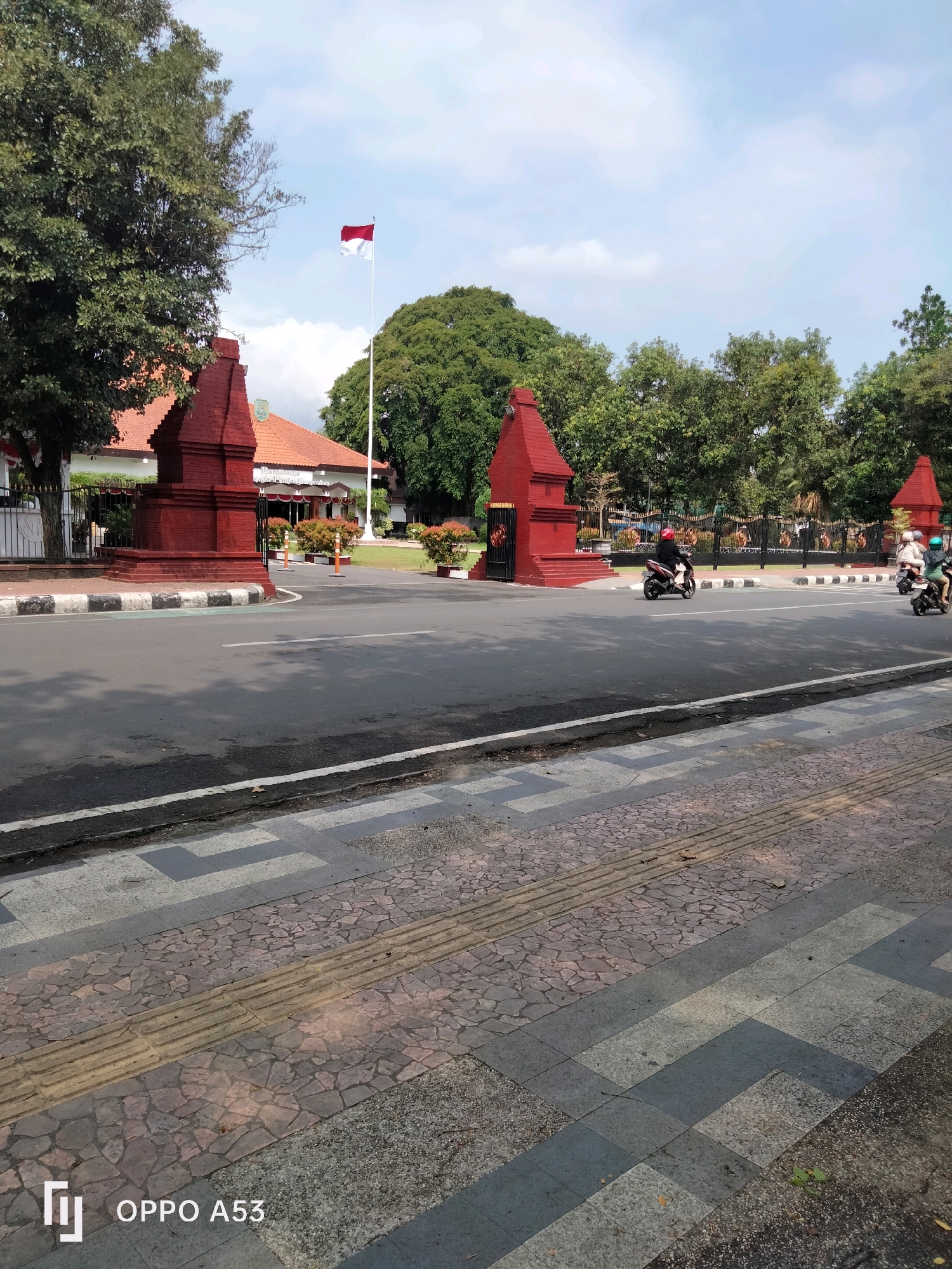 The image shows a street scene in Trenggalek, Indonesia. In the background, there is a building with a red-tiled roof and an Indonesian flag flying from a flagpole. Two red, pyramid-shaped structures are visible on either side of an entrance to the building. The street in the foreground has a pedestrian walkway with tactile paving. Two people on motorcycles are riding on the street. The sky is cloudy.