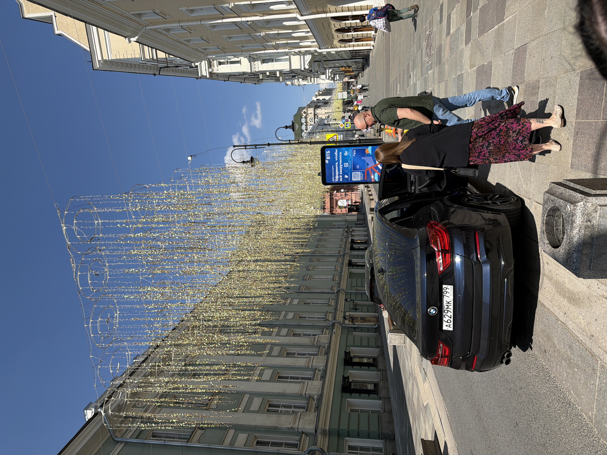 The image captures a street scene in Moscow, Russia, likely a pedestrian area due to the lack of vehicle traffic and the presence of decorative lighting installations. The most prominent feature is a black BMW vehicle parked near a sidewalk. A couple, a man in a green shirt and jeans and a woman in a black top and patterned skirt, are interacting with the car. Adjacent to the car is an advertising display. The background reveals several buildings with a classical architectural style, and there are other people walking along the street further in the distance. The sky is clear and blue, suggesting it's a sunny day. The license plate of the car appears to read "A629MK 799."