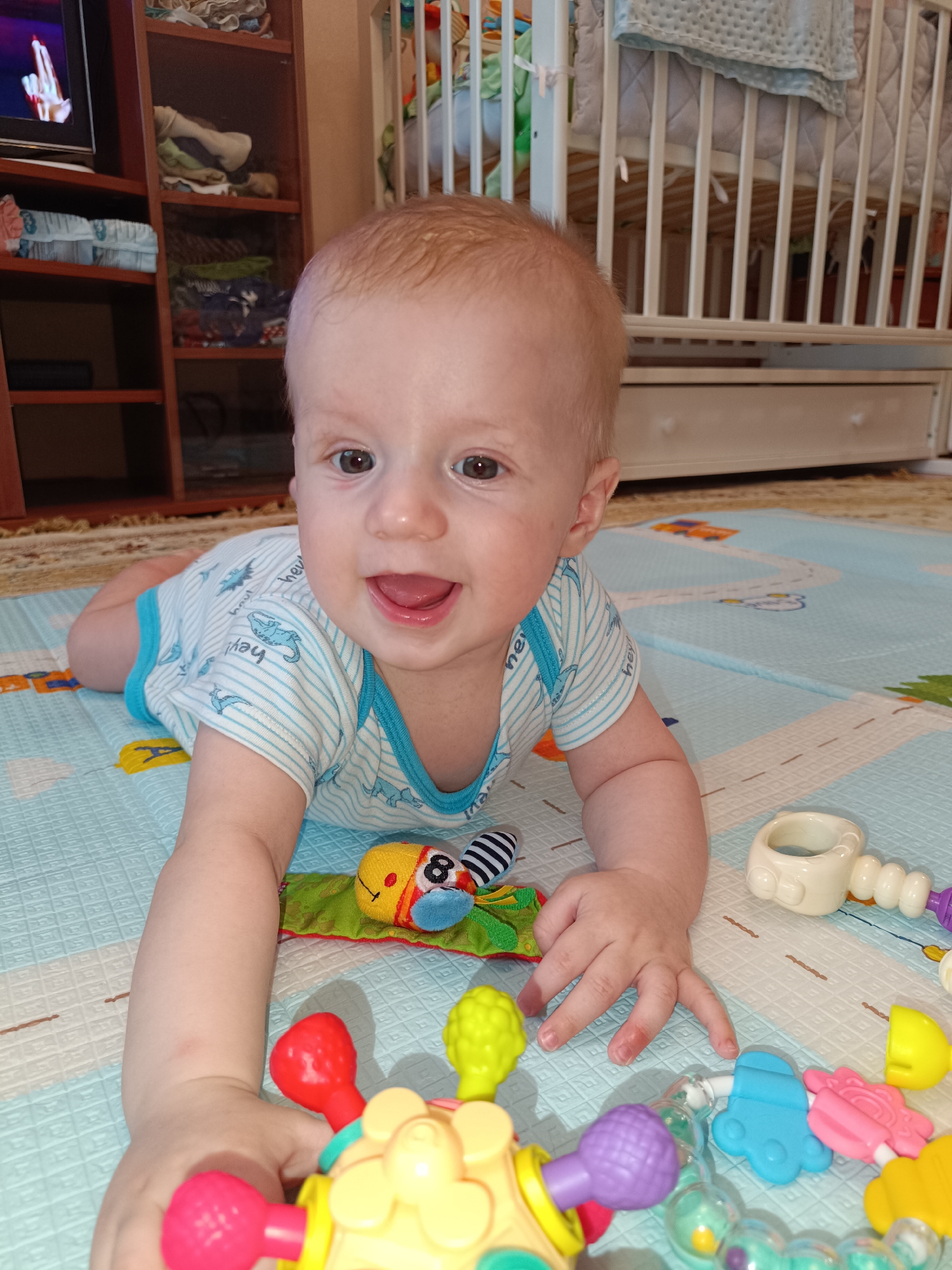 The image captures a close-up, eye-level view of a young, fair-skinned infant lying on its stomach on a blue and white playmat. The infant has light brown hair and is wearing a white and blue striped onesie with a playful pattern of what appear to be cartoonish sea creatures. Its mouth is open in a wide smile, revealing its tongue and some teeth, suggesting the baby is laughing or about to speak. The infant's eyes are open and directed towards the viewer.
The playmat is situated on a patterned rug. Scattered around the infant are various colorful baby toys. To the right of the infant, there is a white, bone-shaped rattle and a multi-colored toy with a yellow body and several red and pink attachments, some of which are circular and bulbous. Other toys, including a blue, paw-shaped teether, a green and yellow caterpillar-like plush toy, and a black and white striped object, are also visible.
In the background, to the left, a portion of a wooden shelf unit is visible, filled with folded clothes and other items. Above the shelves, a television screen displays a bright, blurry image of what appears to be a hand with fingers extended. To the right of the infant, a white crib with vertical slats is prominent, with a light blue knitted blanket draped over its edge. The overall environment suggests a domestic setting, likely a child's play area. There is no indication of the specific location within the city of Tobolsk, Russia.