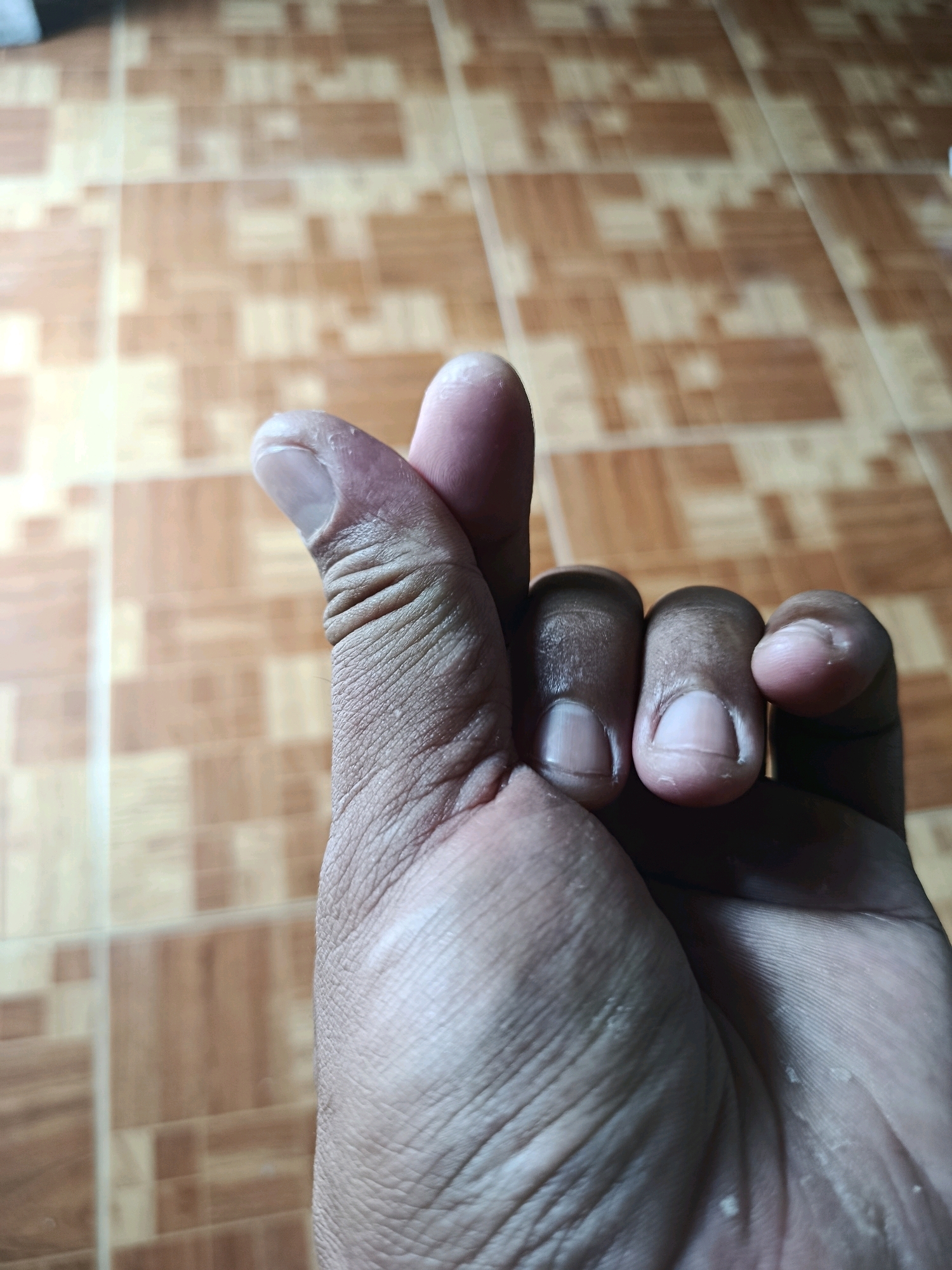 The image is a close-up, ground-level, indoor shot of a person's right hand making a "finger heart" gesture. The hand is positioned in the lower right of the frame, with the thumb and index finger forming a heart shape. The background is a tiled floor with a wood-grain pattern. The lighting appears to be natural, coming from above and slightly to the left, casting subtle shadows. There are no other people or objects visible in the frame. The scene is static and does not depict any activity. No specific location in Binangun, Indonesia, can be identified from the image itself.