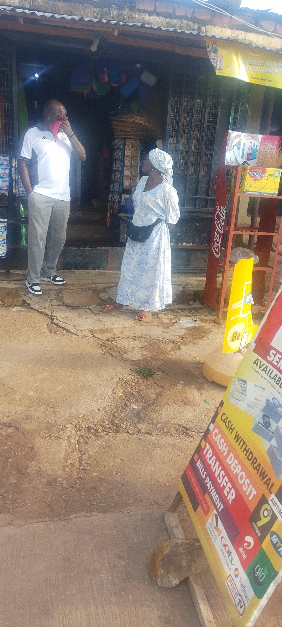 Two individuals are standing outside a storefront in Gura Top, Nigeria. A man on the left, wearing a white polo shirt with gray sleeves and gray pants, is holding a red cloth to his face. A woman on the right, dressed in a long, blue and white patterned dress and a headscarf, is facing away from the camera. A black fanny pack is worn around her waist. In the background, the storefront is filled with various items including bags, packaged goods, and what appear to be brooms. A red Coca-Cola branded shelf unit stands to the right of the storefront, and a yellow advertising sign is placed on the ground in front of it. A larger, colorful sign with text such as "CASH WITHDRAWAL," "CASH DEPOSIT," and "TRANSFER" is leaning against a wooden structure in the foreground, obscuring part of the ground. The ground appears to be a mixture of concrete and dirt, with some patches of sparse green grass. The lighting suggests an overcast or shaded environment.
