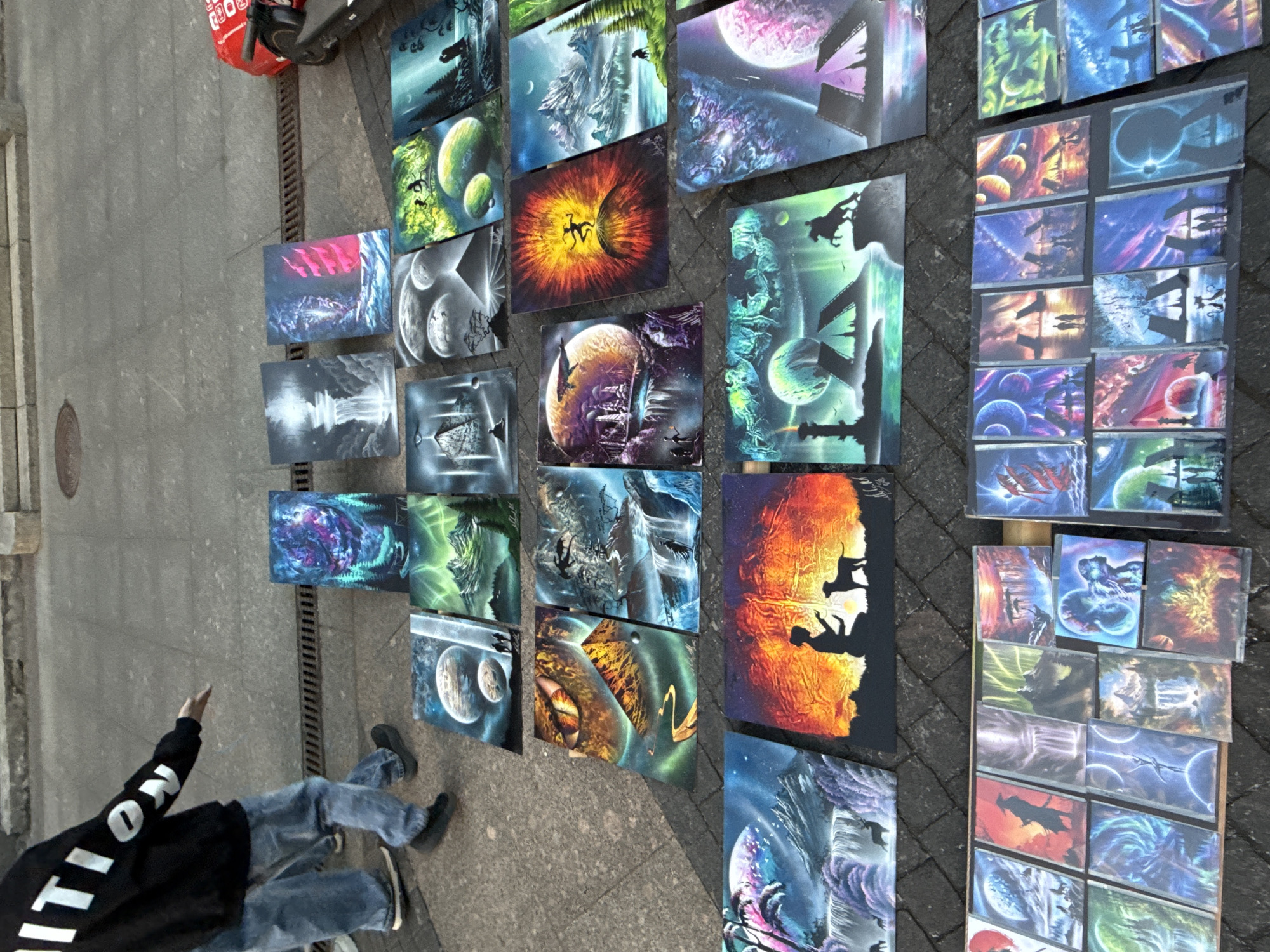The image depicts an outdoor art display on a paved sidewalk in Saint Petersburg, Russia. The primary subjects are numerous spray-painted artworks featuring fantastical, often extraterrestrial, landscapes and scenes. These paintings display vibrant colors and imaginative imagery, including planets, nebulae, alien structures, and silhouetted figures.
A person, likely a vendor or artist, is partially visible in the lower left corner of the image. They are wearing a black hoodie with white lettering on the back, which appears to say "EDITION" or something similar, and blue jeans. The person is sitting or kneeling on the ground, with their right arm extended towards the artwork, possibly gesturing or interacting with a potential customer.
The setting appears to be daytime, with diffuse lighting suggesting an overcast or cloudy day, which is common for Saint Petersburg. The ground is paved with interlocking bricks, and a metal grate or drain runs along the edge of the sidewalk where the art is displayed. A red bag and part of a scooter are visible in the upper left, indicating the urban environment. The overall mood is one of casual commerce and artistic presentation. The artwork itself is the most prominent feature, showcasing a range of creative and detailed depictions of otherworldly scenes.