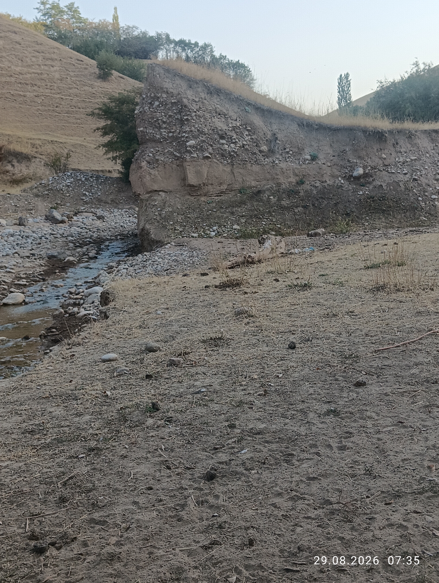 An outdoor daytime full shot captures a dry, rocky stream bed flowing through a barren landscape. The terrain consists of sandy soil, scattered stones, and sparse, dry vegetation. A steep, eroded embankment rises to the left, revealing layers of soil and rock. Trees and shrubs are visible on the distant slopes. The sky is clear and overcast. The image is dated 29.08.2026, 07:35. The location is identified as Ak-Took, Kyrgyzstan.