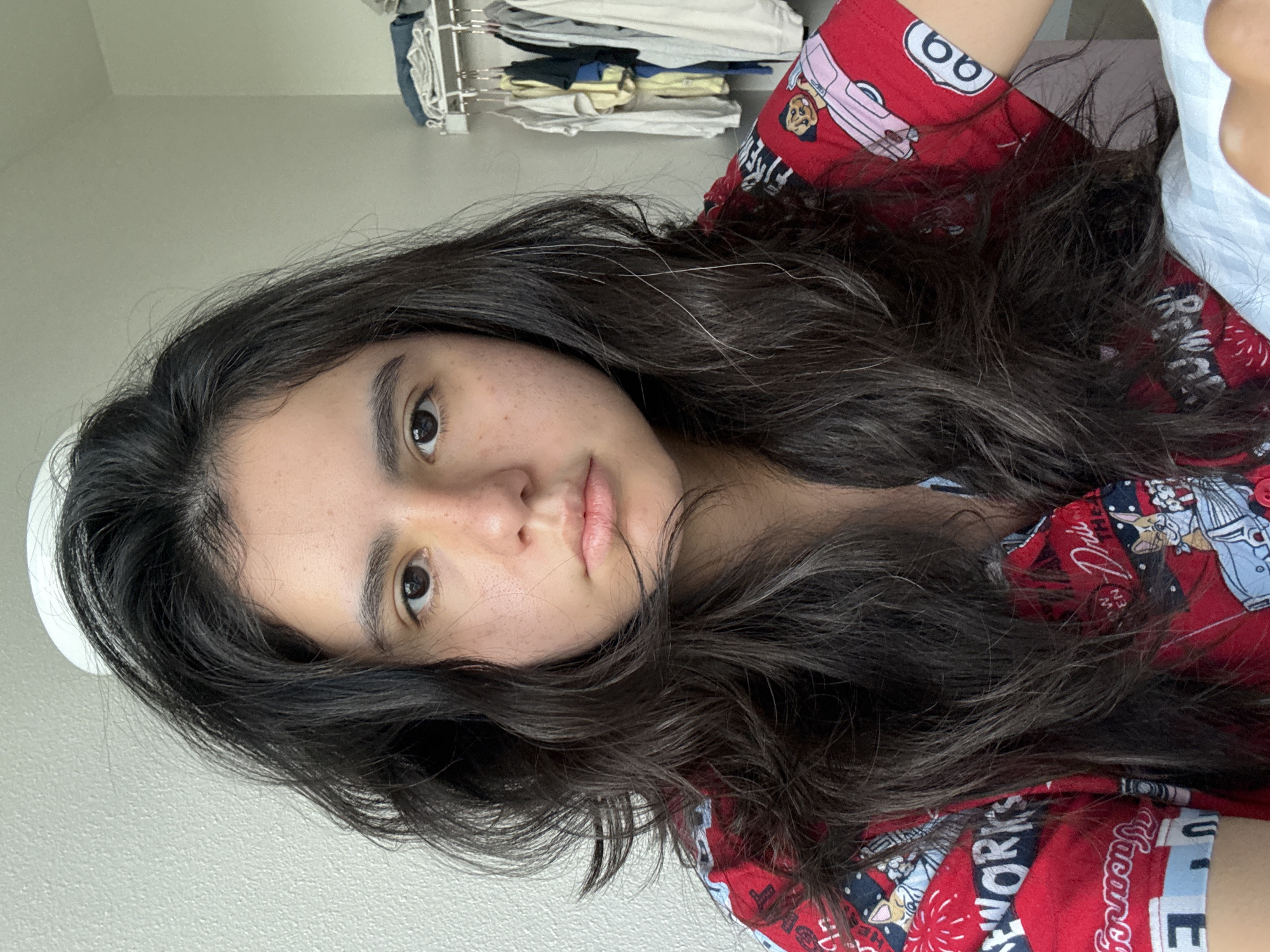 The image presents a close-up, portrait-oriented shot of a young person, likely female, positioned horizontally with their head resting on a surface, looking directly at the camera. The subject has long, dark, wavy hair that flows around their face and extends across their chest. Their complexion is light to medium with noticeable freckles on their cheeks, and they have dark eyes and full lips, maintaining a neutral and calm expression. Individual strands of white or grey hair are visible amidst the dark hair.
The person is wearing a red patterned top featuring various cartoon graphics. Distinct designs include a pink car with a character and the number "66" enclosed in a white circle, as well as illustrations resembling fireworks. Visible text fragments on the shirt include "FIREWORKS," "Drie," and "Fancy."
The background is a light-colored wall, indicating an indoor setting, likely a home environment. Above the subject, a clothing rack with various garments, such as t-shirts and folded fabrics, is visible, suggesting a personal space like a bedroom. The lighting is soft and even, consistent with natural daylight indoors. The overall atmosphere conveyed is relaxed and quiet.
