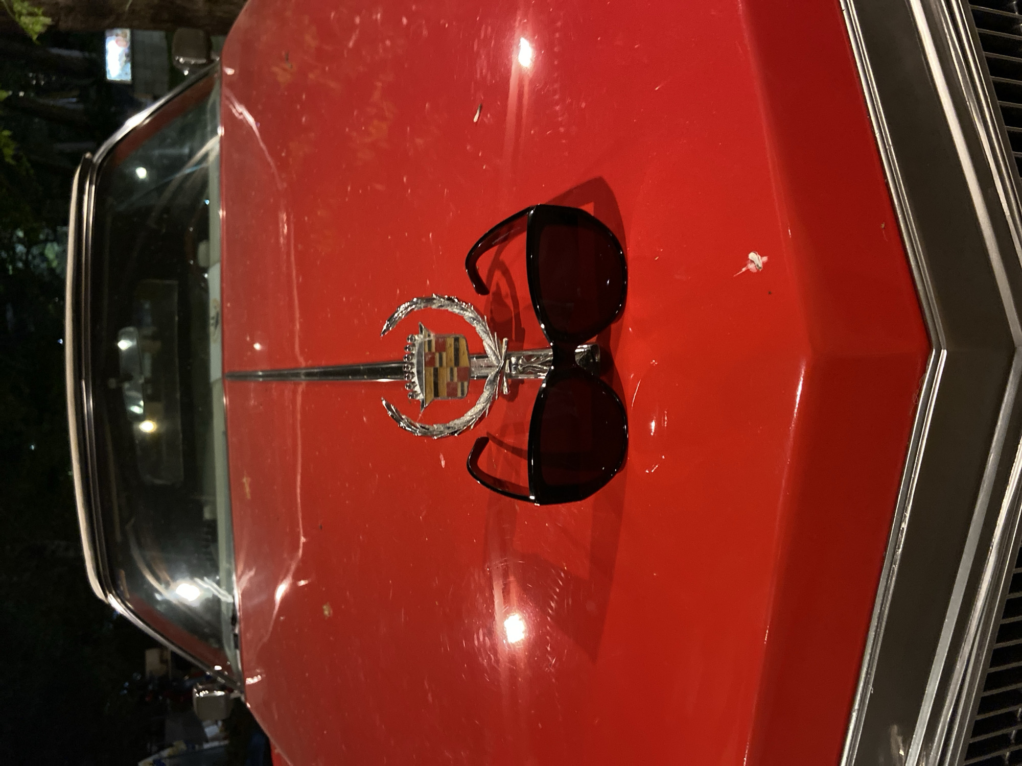 The image shows a red car, possibly a vintage Cadillac, with a hood ornament and a pair of sunglasses resting on the hood. The car is likely parked outdoors, possibly at night, based on the lighting. The location is Budva, Montenegro, but precise placement is impossible.