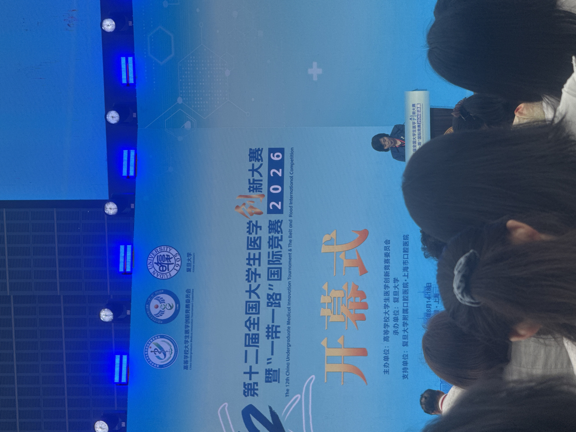 This is a full-shot, eye-level, indoor photograph of a screen displaying event information and a stage area with a speaker, viewed from behind an audience. The screen, which occupies the majority of the frame, is a bright blue with white and orange text. At the top of the screen, there are three circular logos, two on the left and one on the right. Below the logos, the screen displays event titles in Chinese and English. The top line of Chinese text reads "第十二届全国大学生医学创新大赛," and below it, in smaller English text, is "The 12th China Undergraduate Medical Innovation Tournament." To the right of this, a smaller line of Chinese text reads "暨‘一带一路’国际竞赛" and below it in English is "& The Belt and Road International Competition." The year "2026" is prominently displayed in large, bold numbers. Below the event titles, the word "开幕式" (Opening Ceremony) is written in large, bold orange characters. Underneath this, smaller Chinese text indicates the organizing and supporting units. A woman, presumably the speaker, is visible on a stage to the right, standing behind a podium. She is wearing a dark suit jacket and a red necklace and appears to be speaking. The background of the stage area is also blue. The foreground is filled with the backs of the heads of an audience, their hair styled in various ways, obscuring the lower part of the screen and the front of the stage. The lighting appears to be a mix of stage lighting and ambient room light, creating a slightly washed-out effect in some areas. The overall atmosphere is formal, suggesting an academic or competitive event. The mention of Yangpu District, China, in the prompt suggests this event might be held in a university or convention center within that district, likely a location associated with Fudan University given its prominent logo on the screen.