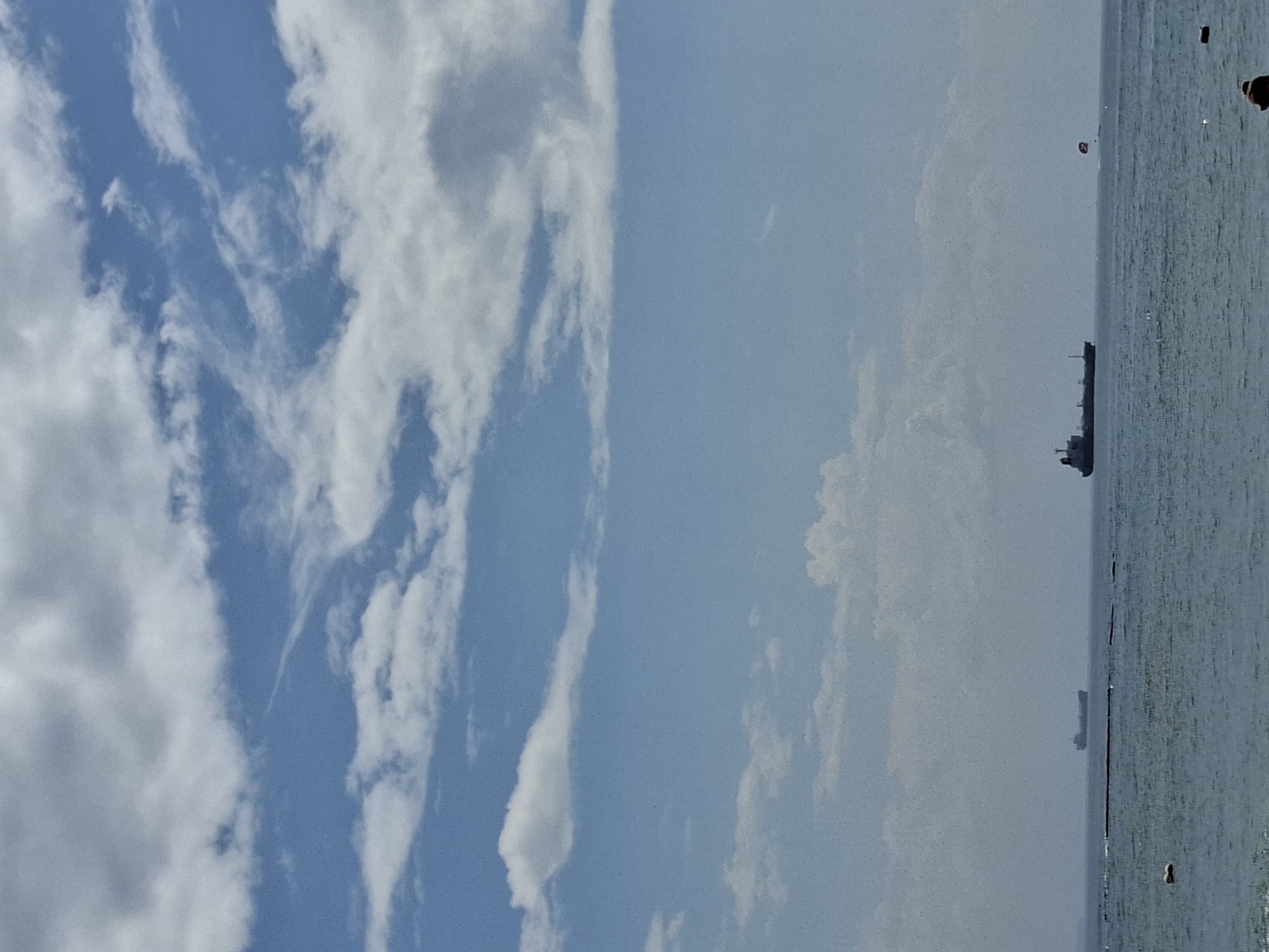 This image captures a serene seascape under a bright, partly cloudy sky, characteristic of a coastal area like Kuta Selatan, Indonesia. The photograph is divided horizontally between the sky and the ocean by a clear horizon line.
The upper half of the image is dominated by an expansive blue sky, filled with scattered white clouds. These clouds vary in form, from wispy and elongated streaks to softer, puffier cumulus-like formations, indicating fair weather conditions. The sky appears bright, suggesting it is daytime.
Below the horizon, the calm ocean stretches across the scene, displaying a light blue-green color. The water surface shows subtle ripples and reflections of the bright sky. In the distance, several maritime objects are visible. A larger, dark-hulled ship with a visible superstructure is prominently positioned on the water towards the center-right, likely a cargo or tanker vessel. Further away, a smaller, fainter ship can be discerned. A red and white object, possibly a buoy or a navigational marker, is also visible floating on the water beyond the larger ship. The very right edge of the image shows indistinct dark shapes that could be distant land, rocks, or artificial structures.
No people are present in the image. The scene depicts a peaceful, clear day, likely late morning or early afternoon, with good visibility. The overall impression is one of tranquil maritime activity. There is no visible text within the photograph.