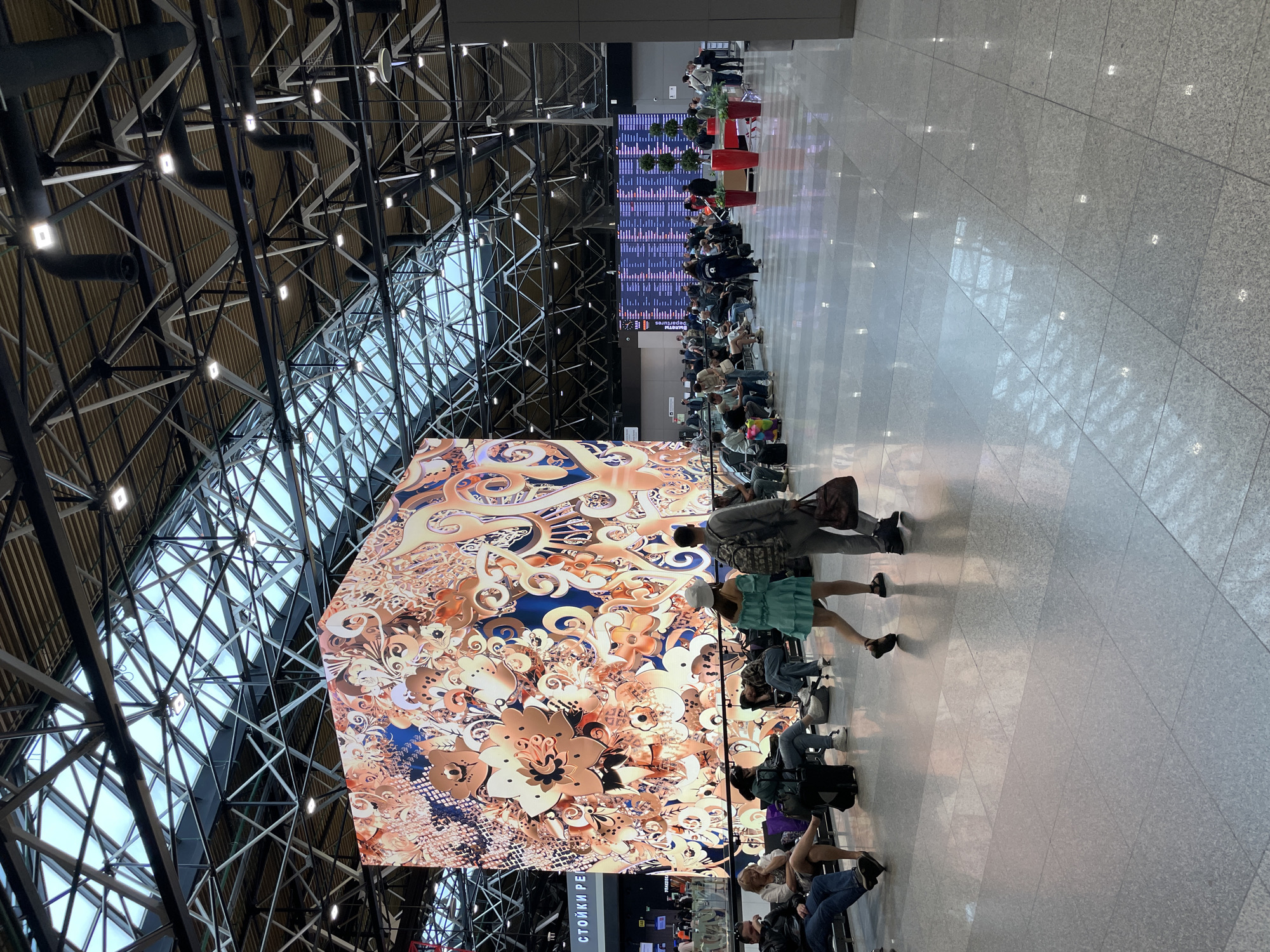 The image depicts the interior of a modern airport terminal, likely located in or near Перепечино, Russia, given the visible signage. The space is vast and open, with a high, industrial-style ceiling featuring a complex network of metal beams and a translucent roof that allows natural light to filter in. Large, bright LED screens dominate the scene. One massive cube-shaped screen displays an intricate, abstract floral pattern in warm orange, gold, and blue tones. In the background, another large screen shows a departure or arrival board with rows of text and numbers.
The floor is polished and reflective, made of large gray tiles. Several groups of people are seated on benches, waiting, with various pieces of luggage around them. In the foreground, two individuals are walking away from the camera. One is a woman in a short, light blue dress and a white bucket hat, and the other is a man carrying a dark brown bag. The overall atmosphere is that of a busy, well-lit transportation hub.