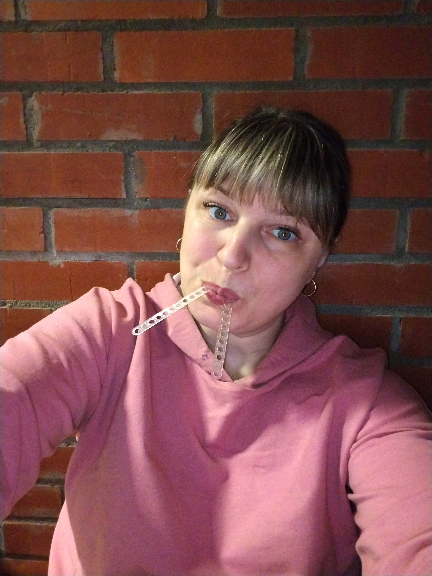 A woman is captured in a close-up portrait, looking directly at the camera with a playful or curious expression. She has light brown or dark blonde hair with bangs covering her forehead and prominent blue-green eyes. Her mouth is pursed around a translucent, segmented plastic object that features small circular holes along its length. She is wearing a pink hooded sweatshirt and small golden hoop earrings. The background consists of a red brick wall with dark mortar, suggesting an indoor setting. There is no visible text in the image. The location is contextualized as Rusanovo, Russia.