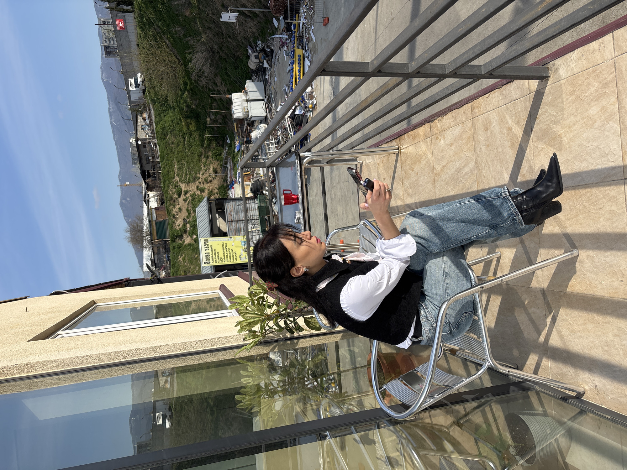 The image depicts a young woman relaxing on a sunlit balcony in Tbilisi, Georgia. She is seated on a modern, chrome-colored metal chair, facing away from the camera, with her legs crossed. She wears a white long-sleeved shirt, a black sleeveless vest, light blue jeans, and pointed-toe black boots. Her dark hair is pulled back, and she holds a smartphone in her right hand, seemingly focused on its screen. A red mug rests on a small table beside her, and a green potted plant is visible on the balcony floor.
The balcony features a tiled floor and a metal railing. Bright sunlight casts sharp, elongated shadows of the railing and the chair onto the floor, suggesting a clear, sunny day, likely late morning or early afternoon.
The background view from the balcony is expansive. Distant mountains rise under a clear blue sky. Directly below the balcony is an area that appears to be an industrial site or junkyard, containing various materials, debris, and structures. Green hillsides frame this area. To the left, a building with large, reflective glass windows mirrors parts of the environment. A yellow sign with Georgian script, "მწვანე აფთიაქი" (translating to "Green Pharmacy"), is visible on a structure in the mid-ground. The overall scene blends natural landscapes with an urban/industrial foreground, observed from a serene vantage point.