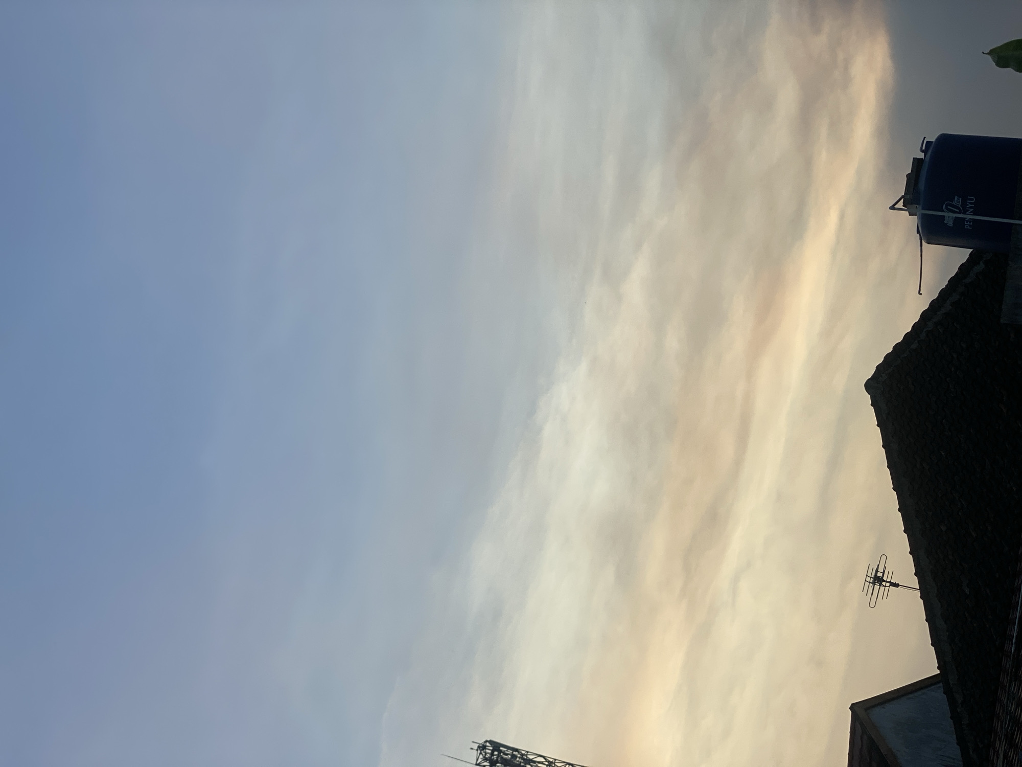 The image captures a skyward view of rooftops and the sky, likely at dusk or dawn, in Banjarbaru, Indonesia. The sky is a gradient of pale blue at the top, transitioning to softer, warmer tones of peach and cream closer to the horizon, with wispy clouds stretched horizontally across. In the foreground, silhouetted against the sky, are the dark, angular shapes of tiled rooftops. On the left, a tall, dark antenna tower rises above a rooftop. Towards the right, a dark blue water tank with white lettering, possibly "PENYU," is visible on top of a roof. The overall scene is tranquil and atmospheric, suggesting an urban or suburban setting bathed in the soft light of early morning or late evening.