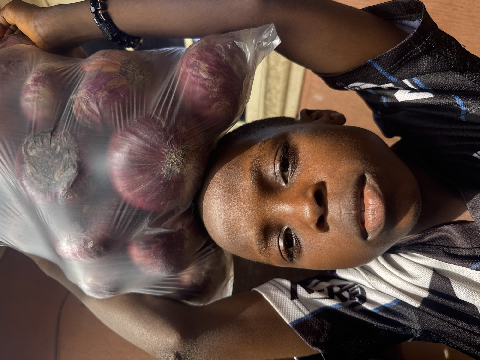 A young person of African descent is holding a plastic bag filled with red onions. The person is wearing a black and white sports jersey and a beaded bracelet. The background is blurred, but appears to be an outdoor urban setting, likely in Onitsha, Nigeria.