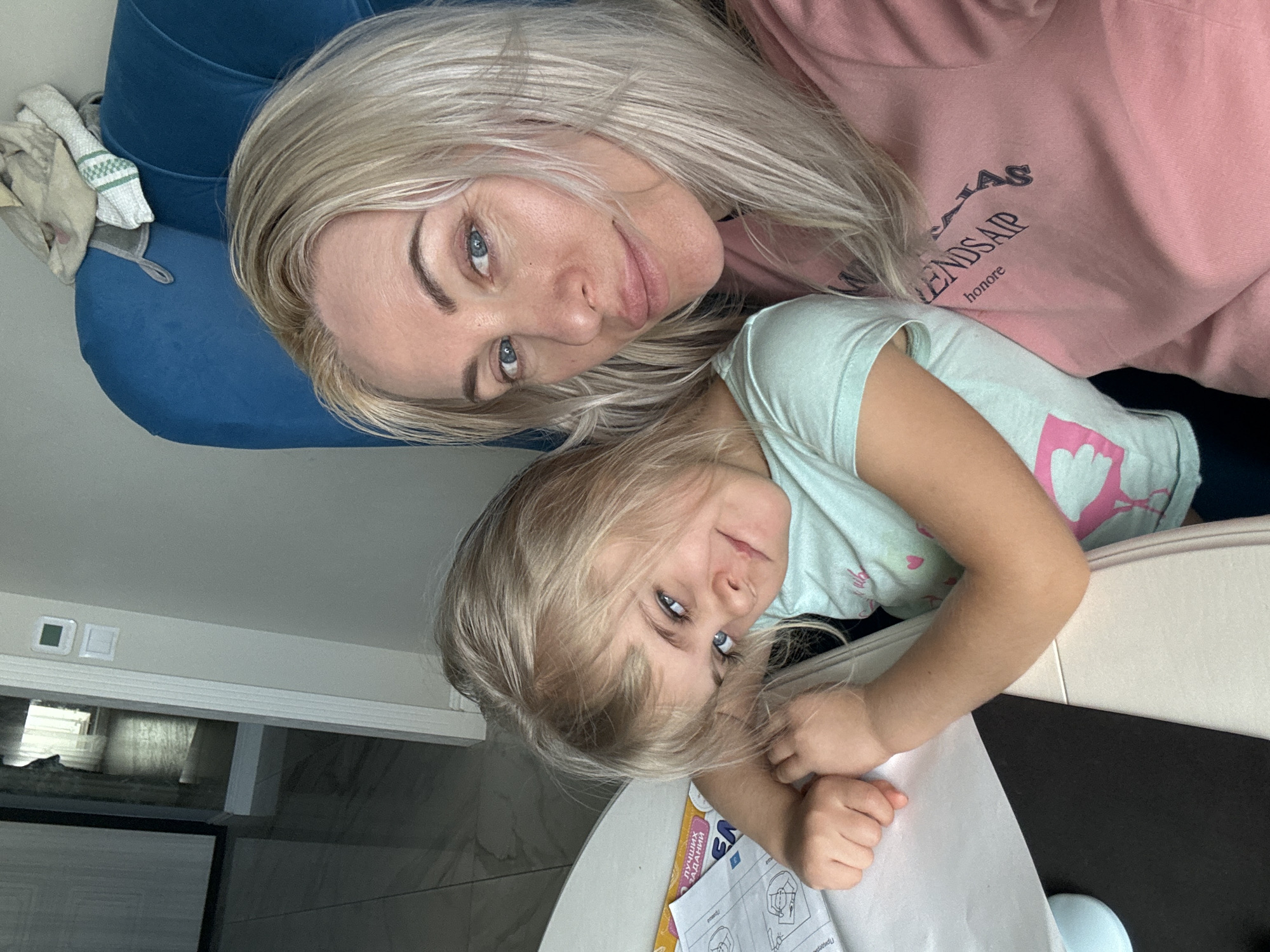 A medium shot, taken indoors, presents a woman and a young child. The woman, positioned at the top, has blonde hair and blue eyes, and is wearing a pink sweatshirt. The child, below her, also has blonde hair and blue eyes, wearing a light blue t-shirt with a pink heart. They appear to be posing closely together, possibly taking a selfie. Visible in the background are a blue cushion, a white surface, a document with diagrams, and what appears to be part of an interior space with tiled floors. The location is unknown.