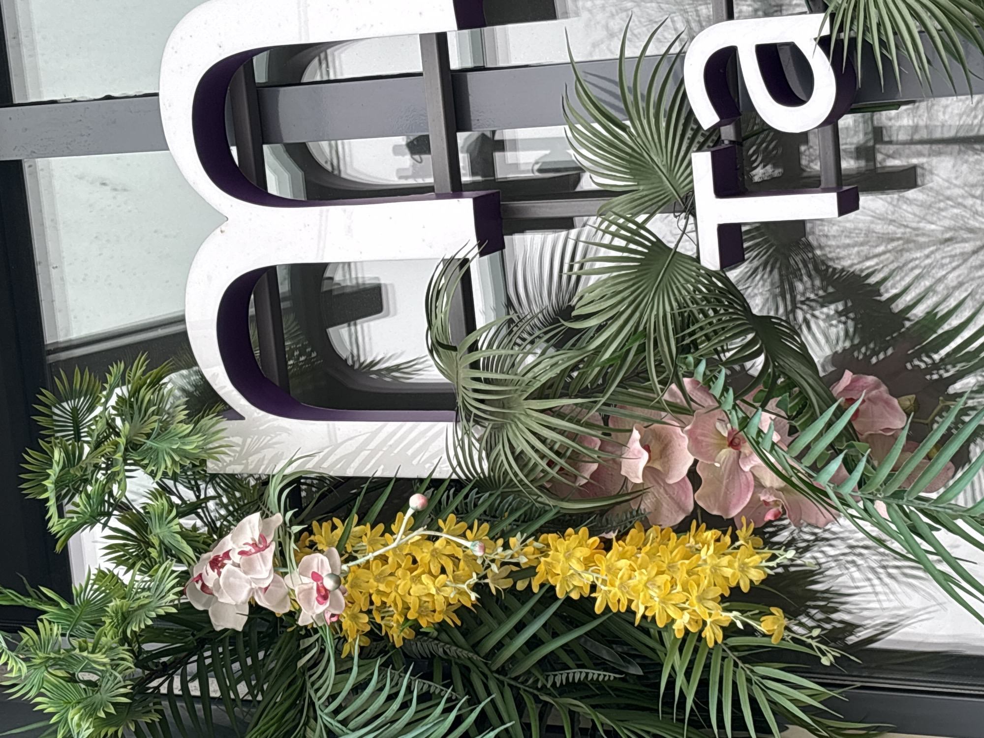 A medium-angle shot shows floral arrangements surrounding a white sign with purple trim that reads "E". The sign is mounted on a dark gray structure. The floral arrangements include a variety of artificial plants and flowers, including yellow and pink orchids, palm leaves, and ferns. The sign is partially obscured by the floral arrangements. The location is not discernible from the image. There are no people visible.