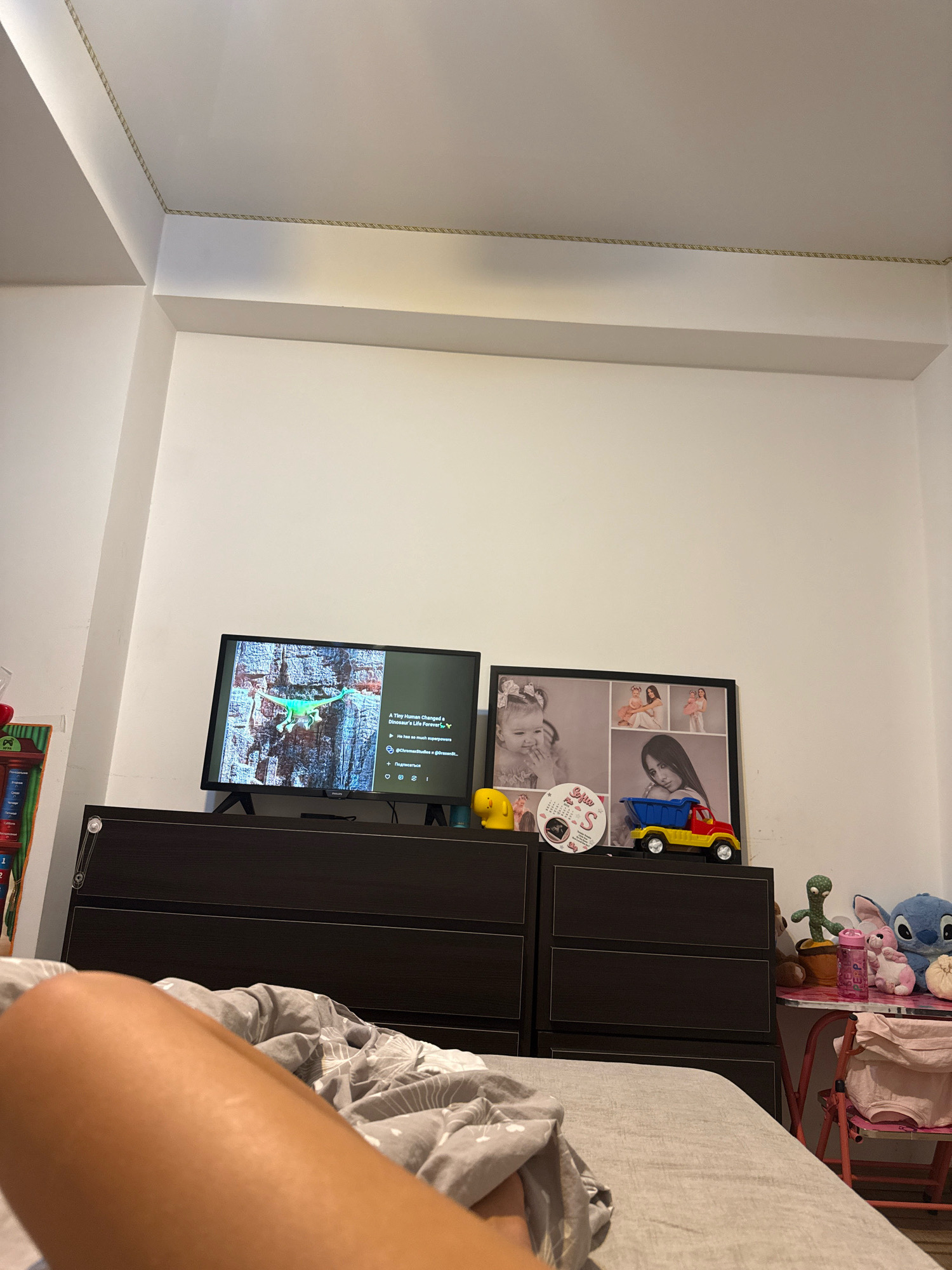 A low-angle indoor shot shows a dresser with a television and a framed collage of photos. The television displays a video of a green dinosaur on a rocky surface. The photo collage features portraits of young children and a woman, with a circular decorative item in the lower left corner. A small yellow toy figure and a toy truck are placed on top of the dresser. To the right, a small table holds stuffed animals, including a cactus, a rabbit, and Stitch, along with a pink water bottle. The overall setting appears to be a bedroom, with a person's leg visible in the foreground, covered by a gray blanket. The walls are white and there is decorative rope molding along the ceiling. There is no information in the image to suggest a specific location in Chișinău, Moldova.