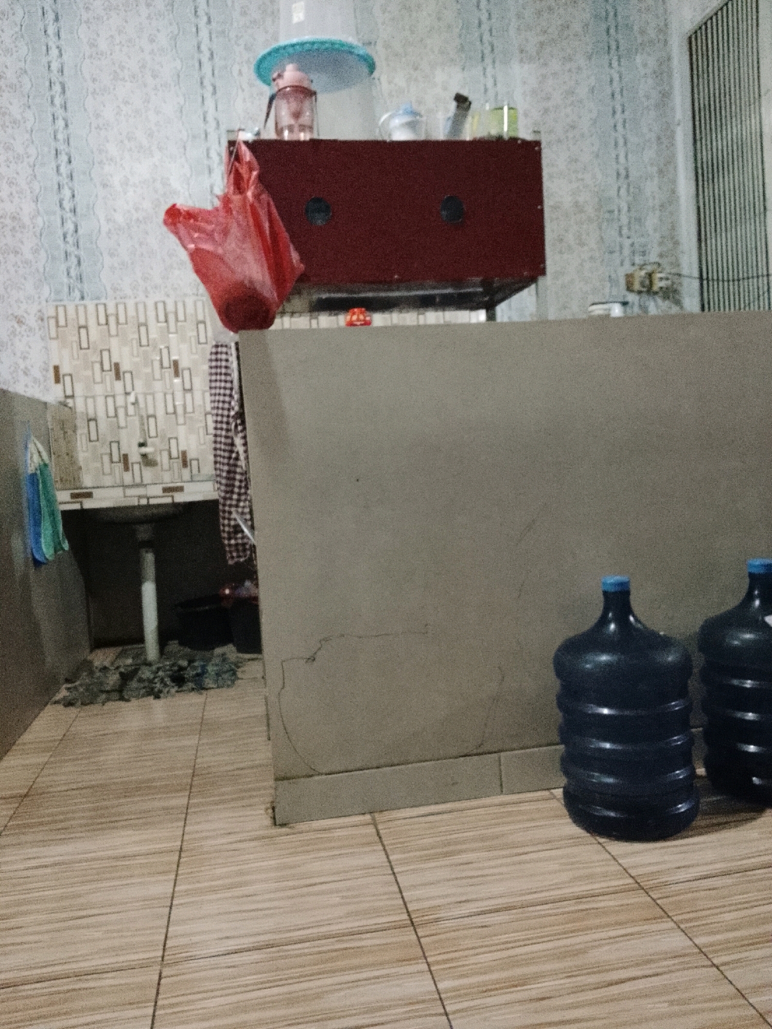 The image depicts an interior scene, likely a kitchen or utility area, with a tiled floor and patterned wallpaper. In the foreground, two large, dark blue plastic water jugs with blue caps are visible. A textured, beige wall extends across the middle of the frame, separating the water jugs from the left side of the image. On the left, a stainless steel sink is partially visible, with a white pipe extending downwards. Above the sink, a tiled backsplash features a repeating pattern of rectangular shapes. A red plastic bag hangs from a hook near the sink. In the background, a dark red cabinet with circular elements is situated. The overall scene is dimly lit and appears to be a functional space. The prompt mentions the city of Padang Dalom, Indonesia, but there are no specific visual cues in the image to confirm this location.