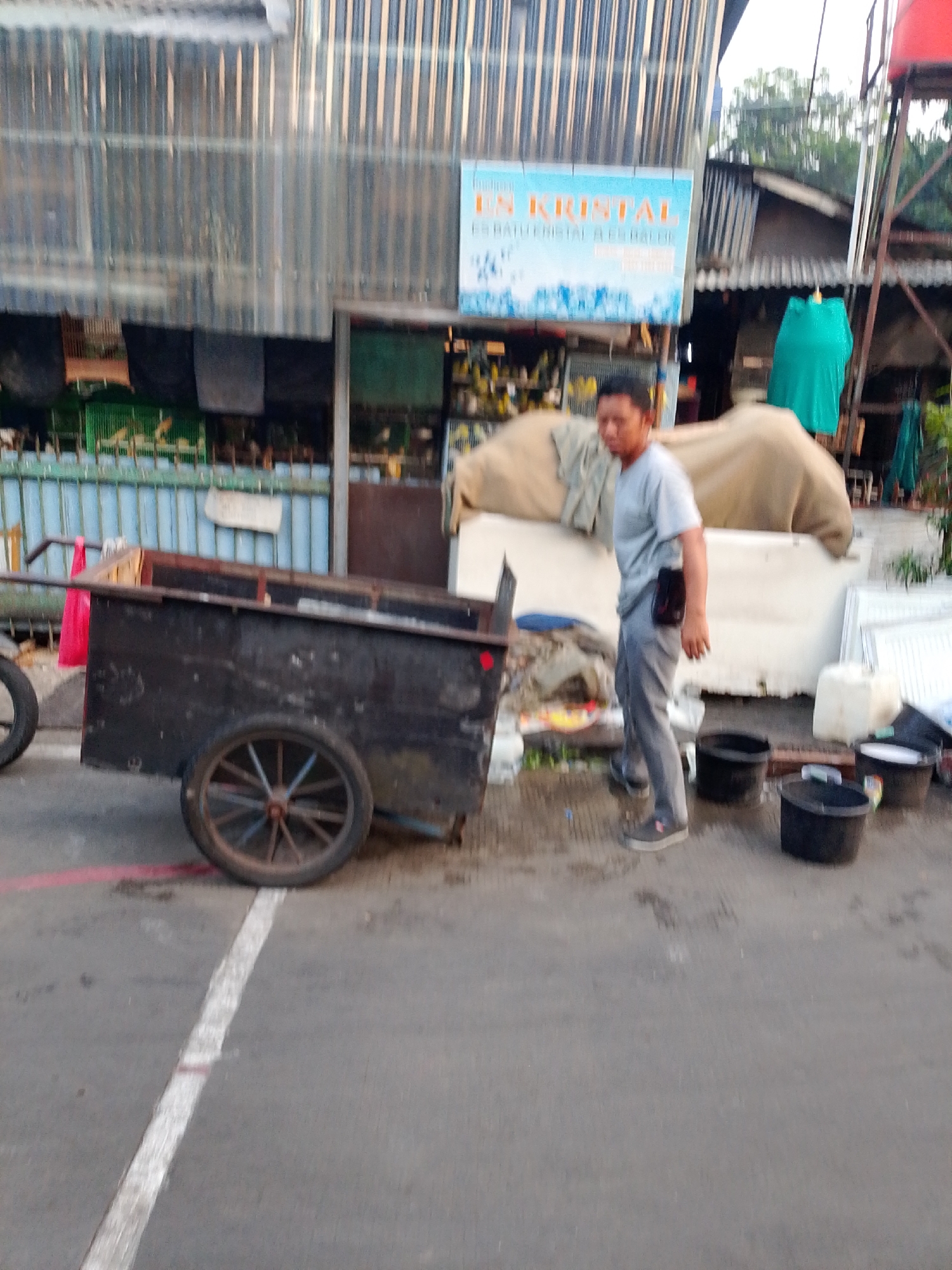 The image depicts a street scene in East Jakarta, Indonesia, with a man standing next to a black wheeled cart. The cart has wooden sides and a metal frame, and a red bag is tied to its handle. The man, wearing a grey t-shirt and grey pants, has a black pouch attached to his waist. In the background, a building with corrugated metal siding is visible, along with various stalls and what appear to be bird cages. Several black buckets are placed on the ground near the man and cart. The ground is wet, suggesting recent rain. A blue sign with Indonesian text, possibly advertising "ES KRISTAL" (ice crystal), is mounted on the building. The overall impression is that of a bustling urban environment with commercial activity.
