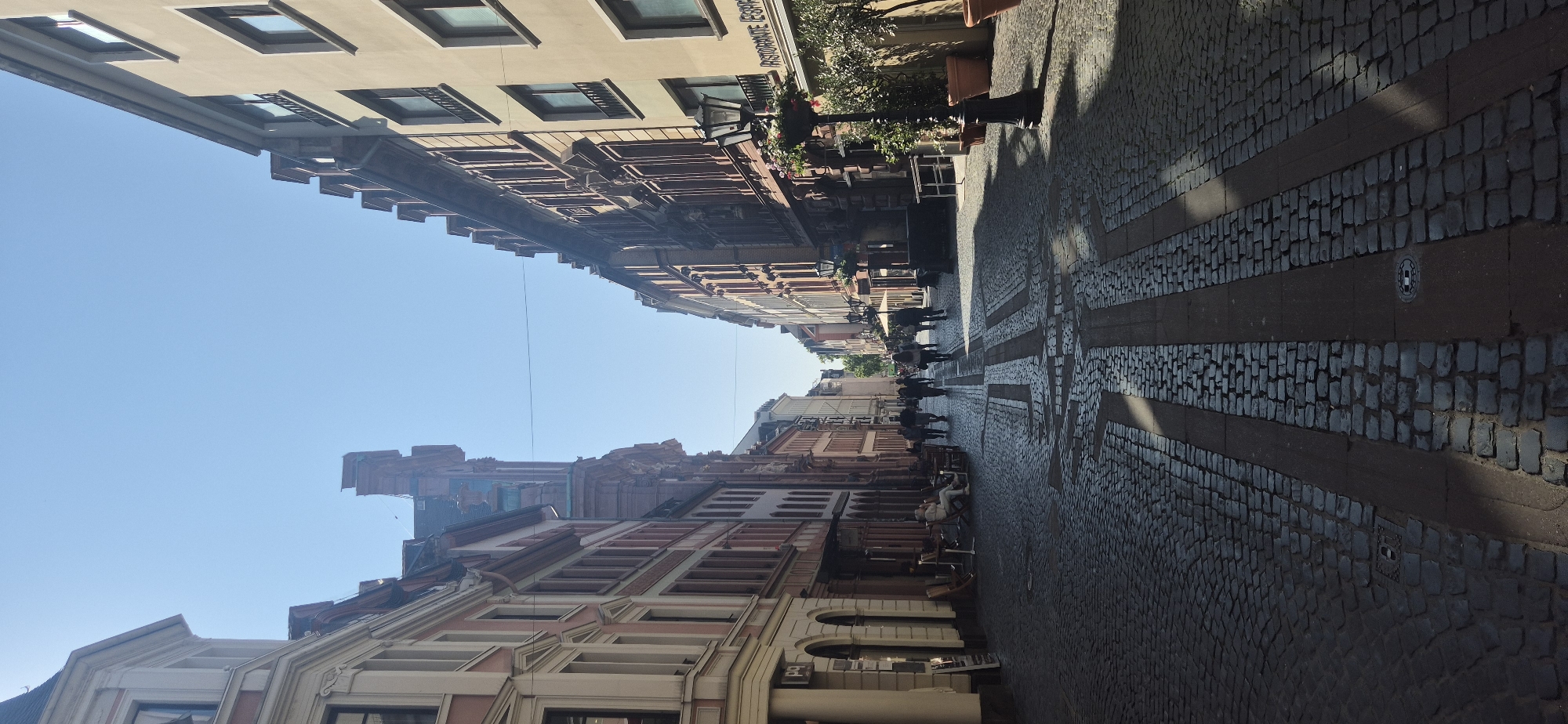 The image depicts a narrow cobblestone street lined with historic buildings in Mainz, Germany. The street is a pedestrian zone with several people walking and seated at outdoor cafes. On the left, there are cream-colored buildings with red accents and arched doorways. On the right, there are buildings with reddish-brown facades, featuring numerous windows and decorative elements. A black lamppost with hanging flowers stands on the right side of the street, near a restaurant with a sign that partially reads "Ristorante Espace". The cobblestone pavement has a pattern of darker stones forming geometric shapes. The sky is clear and blue, indicating a sunny day. The perspective of the image is looking down the street, creating a sense of depth.