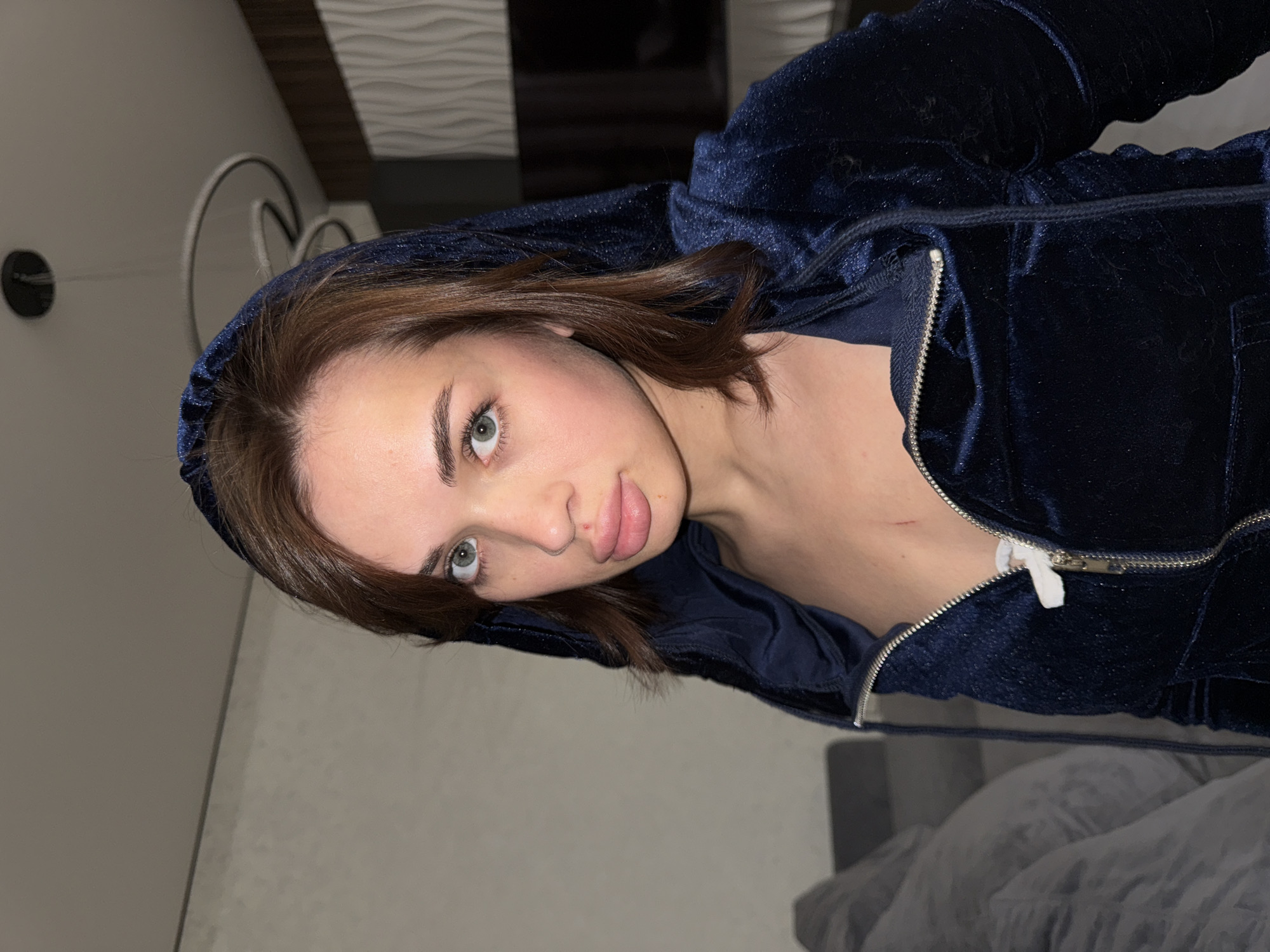 A woman with brown hair and light green eyes is wearing a dark blue velvet hooded sweatshirt. She is indoors in a dimly lit room. The location is inferred as Лобаново, Russia. A partial view of a wall, decorative fixture, and sofa are also visible.