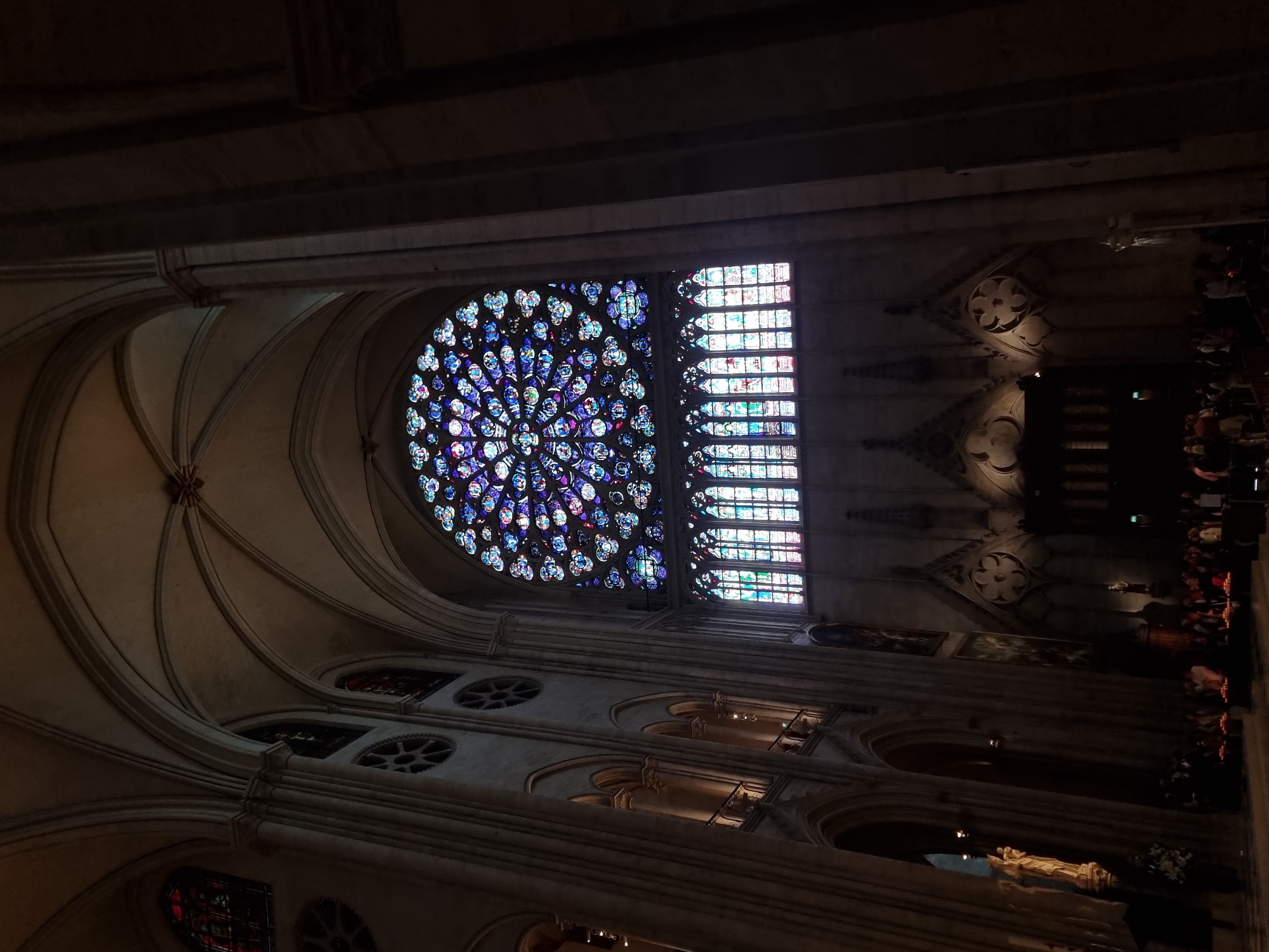 The image shows the interior of Notre Dame Cathedral in Paris, France. The frame is dominated by a large, ornate stained-glass rose window and a rectangular window to its right, both displaying vibrant blues, purples, reds, and greens. Architectural elements like ribbed vaults, arches, and columns are visible, made of light-colored stone. Along the right side of the image, blurred figures indicate an area where people are seated in rows. A statue is visible near the bottom of the frame. Overall, the scene is dimly lit.
