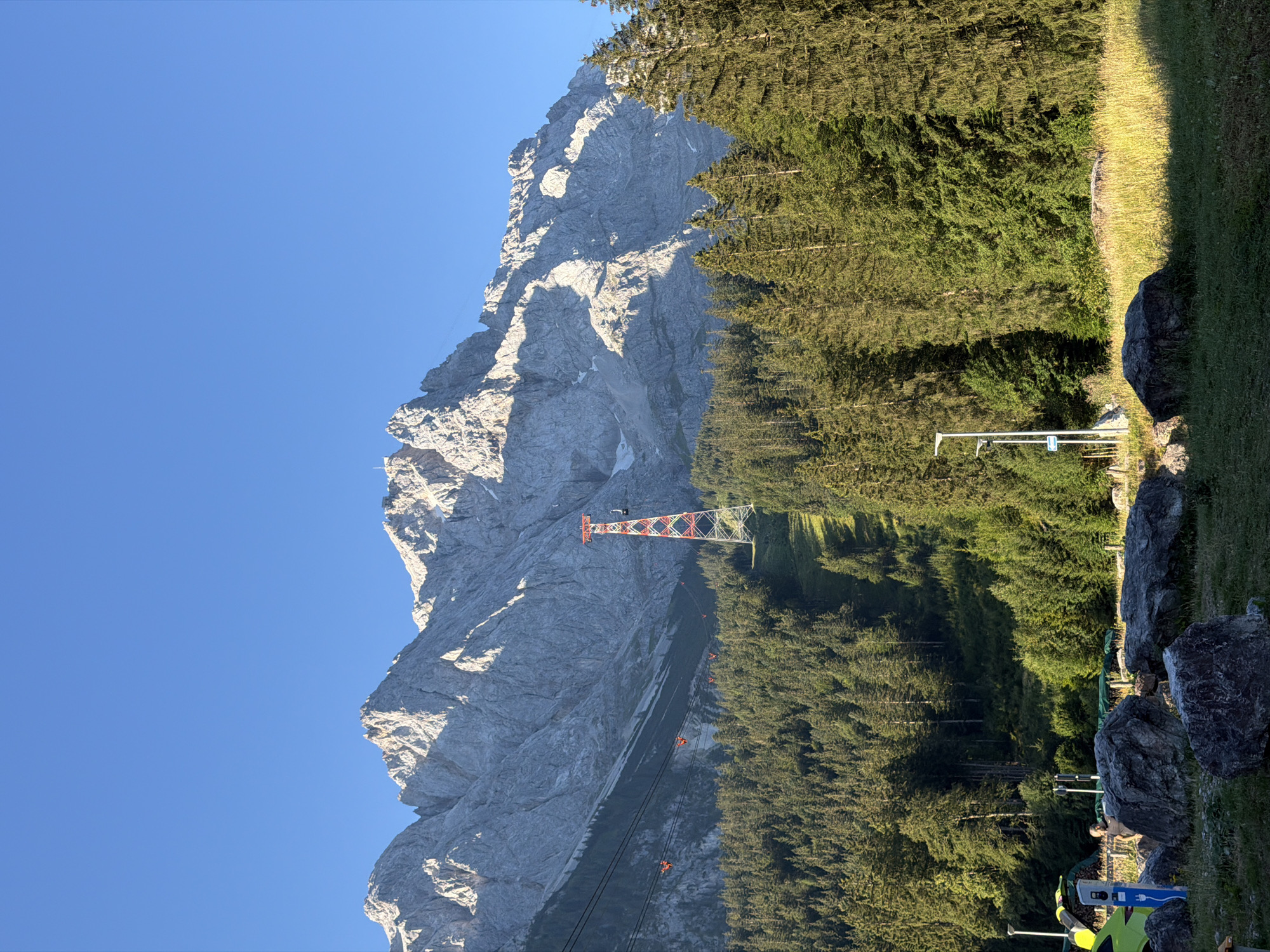 This is a photograph of the Zugspitze cable car in Grainau, Germany. The image shows the steep, rocky slopes of the Zugspitze mountain, covered in dense evergreen forests. A red and white cable car pylon stands tall in the middle of the frame, with the cable cars suspended from wires stretching across the mountainside. In the foreground, large rocks and grassy areas are visible, hinting at a recreational area or parking lot. The sky is clear and blue, indicating a sunny day. The overall scene evokes a sense of natural beauty and outdoor adventure.