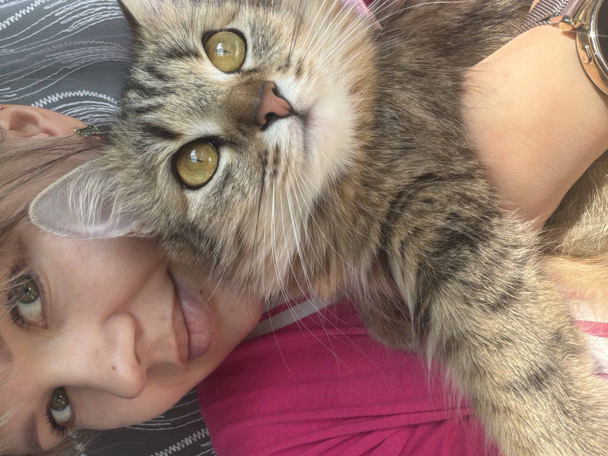 The image shows a light-skinned person with blonde hair, wearing a pink shirt, in close proximity to a tabby cat. The person is lying down, with a portion of their face visible. The cat is positioned partially on the person's body, with its face prominently featured. A wrist wearing a watch is also visible. The background features a gray and white patterned surface. The location is unknown.