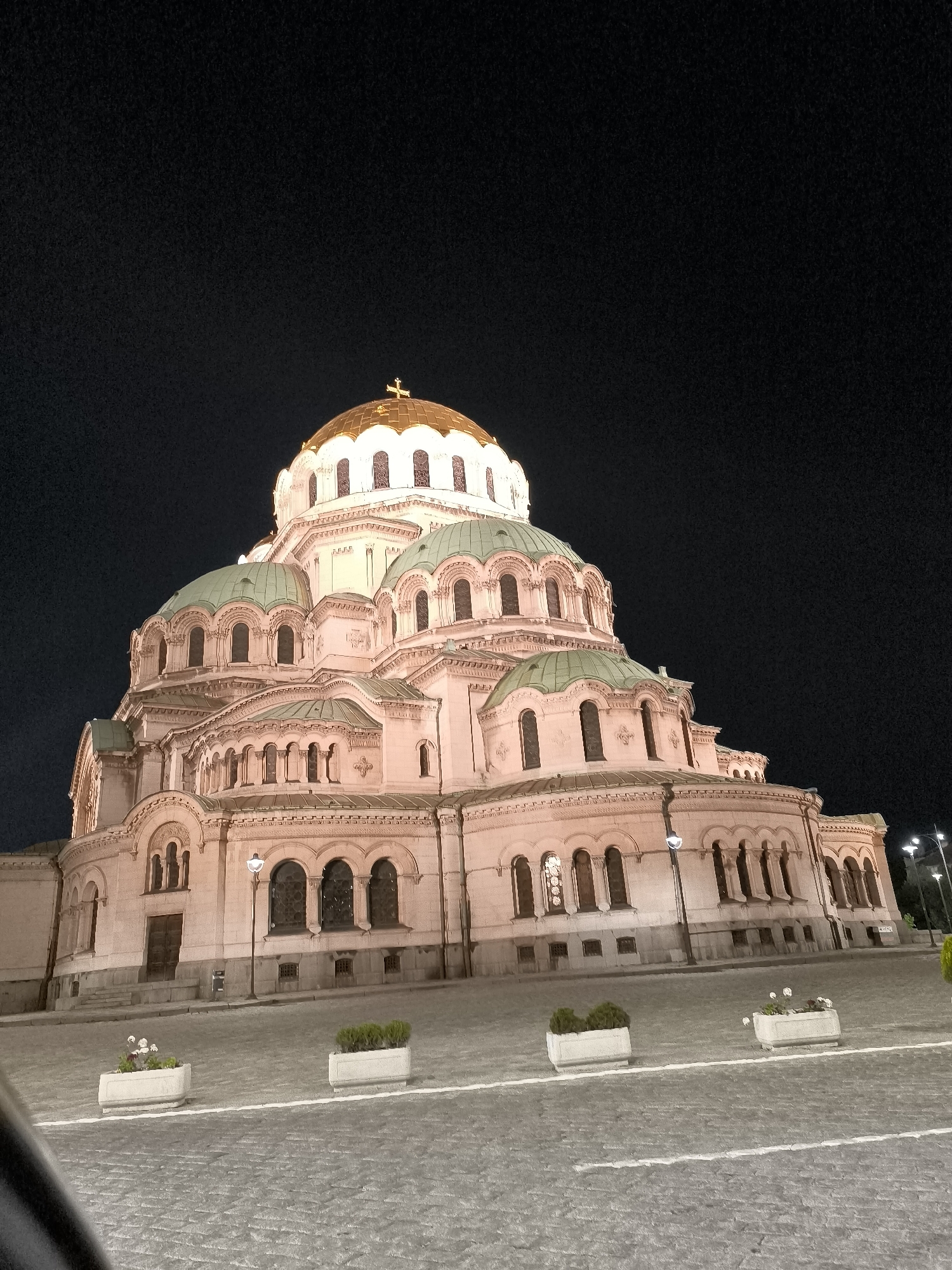 The image displays the majestic Alexander Nevsky Cathedral in Sofia, Bulgaria, illuminated at night. The grand structure dominates the frame, showcasing its distinctive Neo-Byzantine architecture against a dark, starless sky.
The cathedral features a large, prominent central dome with a golden or bronze finish, topped by a brightly lit cross. Numerous smaller domes with a greenish-turquoise patina flank the central one, contributing to the building's impressive silhouette. The stone facade of the cathedral is a warm, light pinkish-beige, softly glowing under artificial illumination which highlights its intricate details, including multiple arched windows, cornices, and decorative elements.
In the foreground, a cobbled plaza or street extends from the bottom of the frame towards the cathedral. Faint white lines are visible on the cobblestones. Several rectangular planters, containing small green shrubs or white flowers, are arranged on the ground. To the far right, distant streetlights can be seen, confirming the nighttime setting. There are no people visible in the photograph, and no discernible text is present. The clear night sky suggests fair weather. The overall impression is one of serene architectural grandeur, with the cathedral standing out as a magnificent beacon in the darkness.