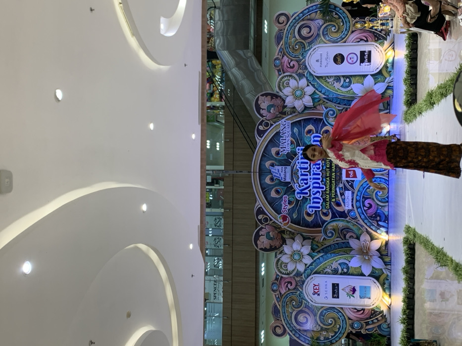 The image shows a fashion show taking place in the Malang Town Square in Malang, Indonesia, located at coordinates -7.957 latitude and 112.619 longitude. A young woman wearing a traditional Indonesian outfit is walking down a runway, smiling and waving her hands. The background is a large colorful stage with the words "Kartika Inspirasi" written on it, along with the logo of the Malang Town Square and several sponsors. The runway is lined with green plants. The scene takes place inside a shopping mall, with a white ceiling and an escalator visible in the background. Some members of the audience are seated in the foreground.