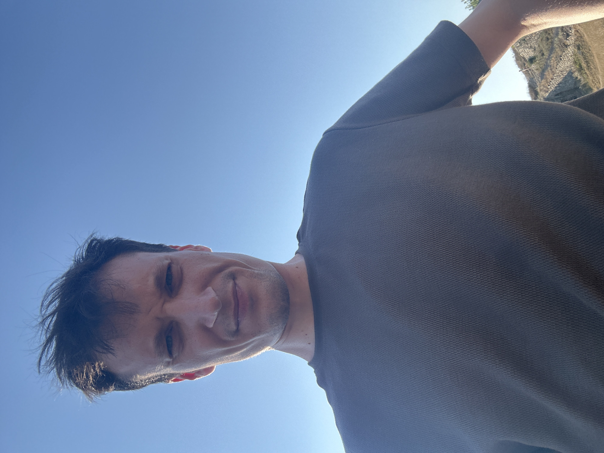 The image is a selfie of a man against a clear blue sky. He is wearing a dark gray t-shirt and his head is tilted slightly back, with his gaze directed towards the camera. His hair is dark and appears windswept. The lighting suggests it is daytime, possibly late afternoon due to the low angle of the sun creating a rim light effect around his face and hair. The lower part of the image shows a slightly blurred background with a rocky, uneven terrain and a few sparse plants, hinting at an outdoor, possibly elevated location. Without specific landmarks visible, the location in Kamianets-Podilskyi, Ukraine cannot be confirmed.