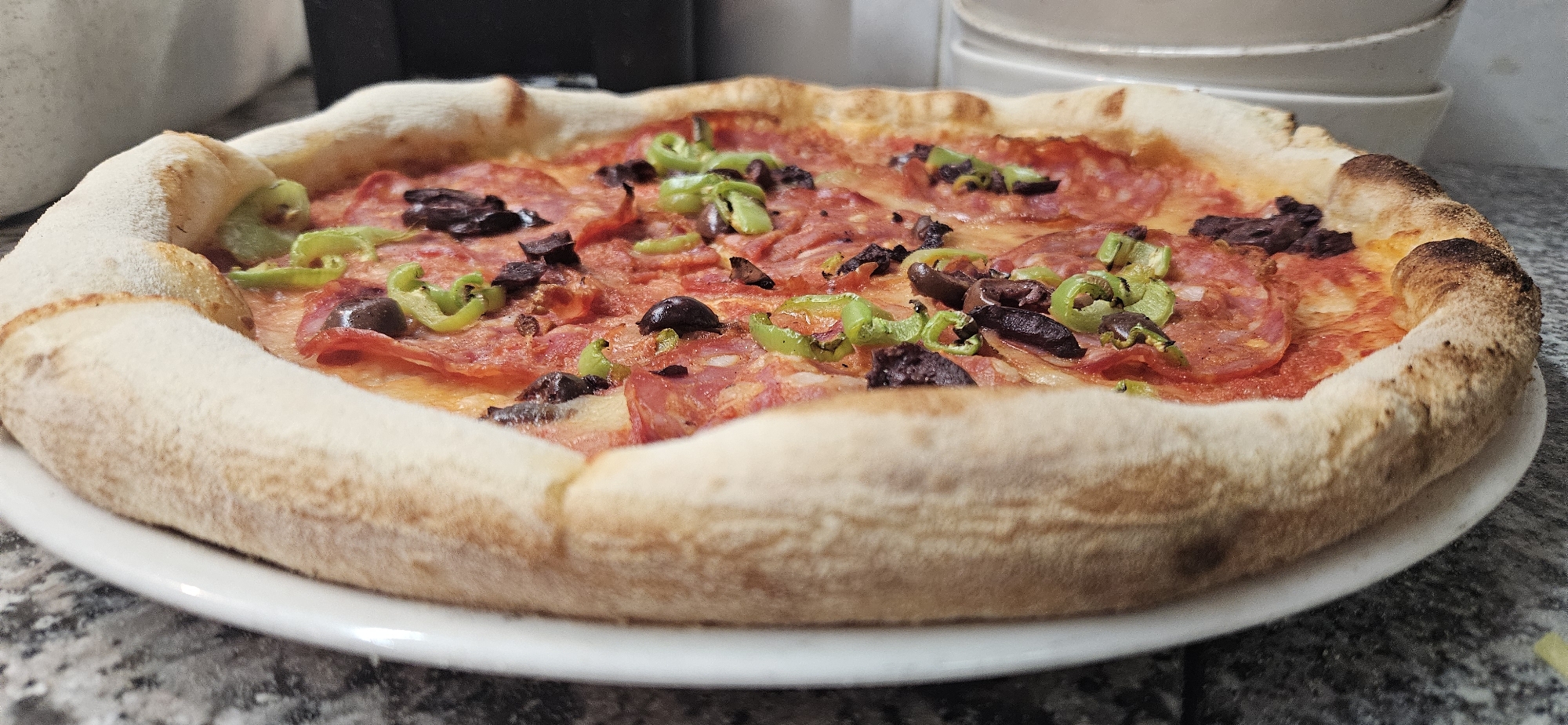 A close-up shot shows a pizza on a white plate set on a grey countertop with a marbled pattern. The pizza has a thick crust that is lightly browned. The toppings include salami, slices of green peppers, and what appears to be olives or a dark topping. A stack of white bowls is partially visible in the background. The location is indeterminate.