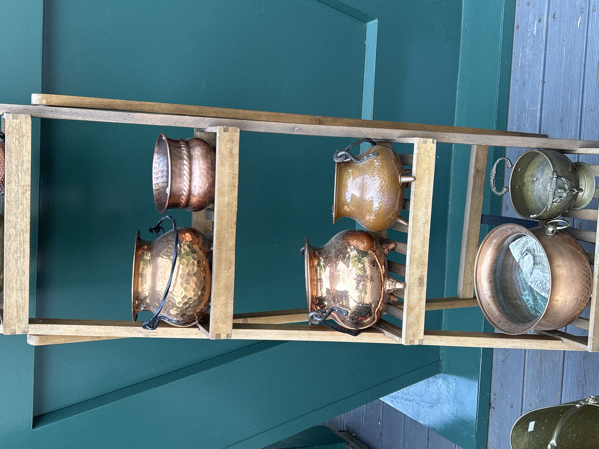 The image shows a wooden rack holding a collection of copper and brass pots and bowls. The rack is leaned against a green wall on the left and blue-grey weathered wood on the right. The metal pots are of varying sizes and shapes, with some having decorative details. The setting is outdoors, but the exact location is not apparent from the image.