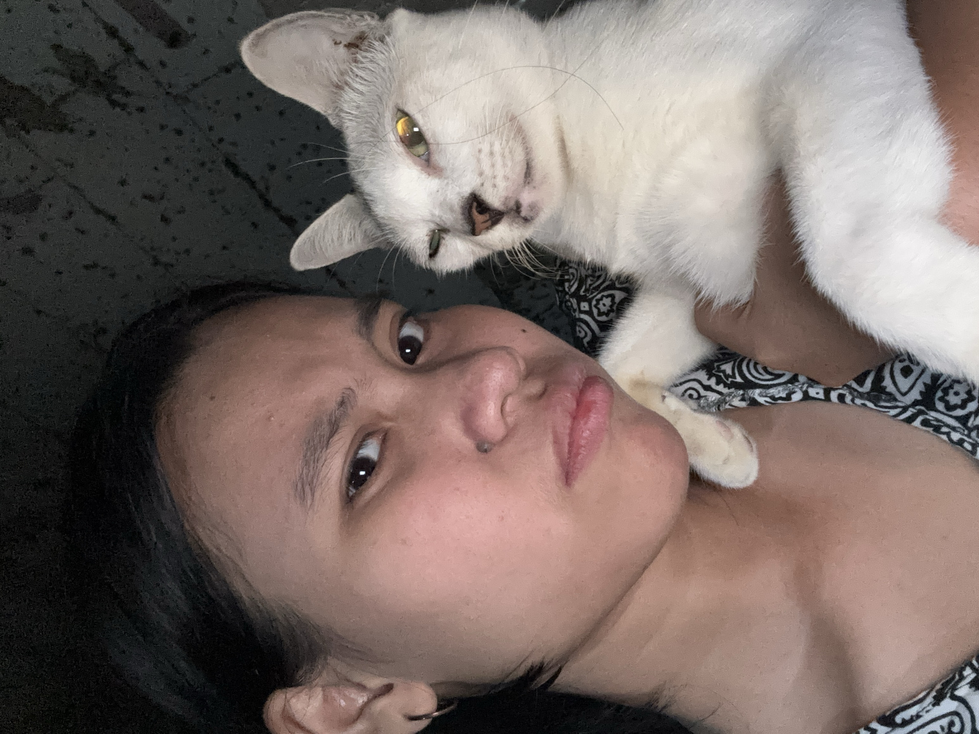 A selfie taken at night shows a young woman holding a white cat in East Jakarta, Indonesia. The woman, positioned on the left side of the frame, has her face turned towards the camera with a displeased expression, her mouth slightly pursed. She has dark hair pulled back and is wearing a hoop earring in her left ear. The cat, on the right, is white with light grey markings, and appears to be winking, with its head tilted. The background is dark and indistinct, suggesting an indoor setting.