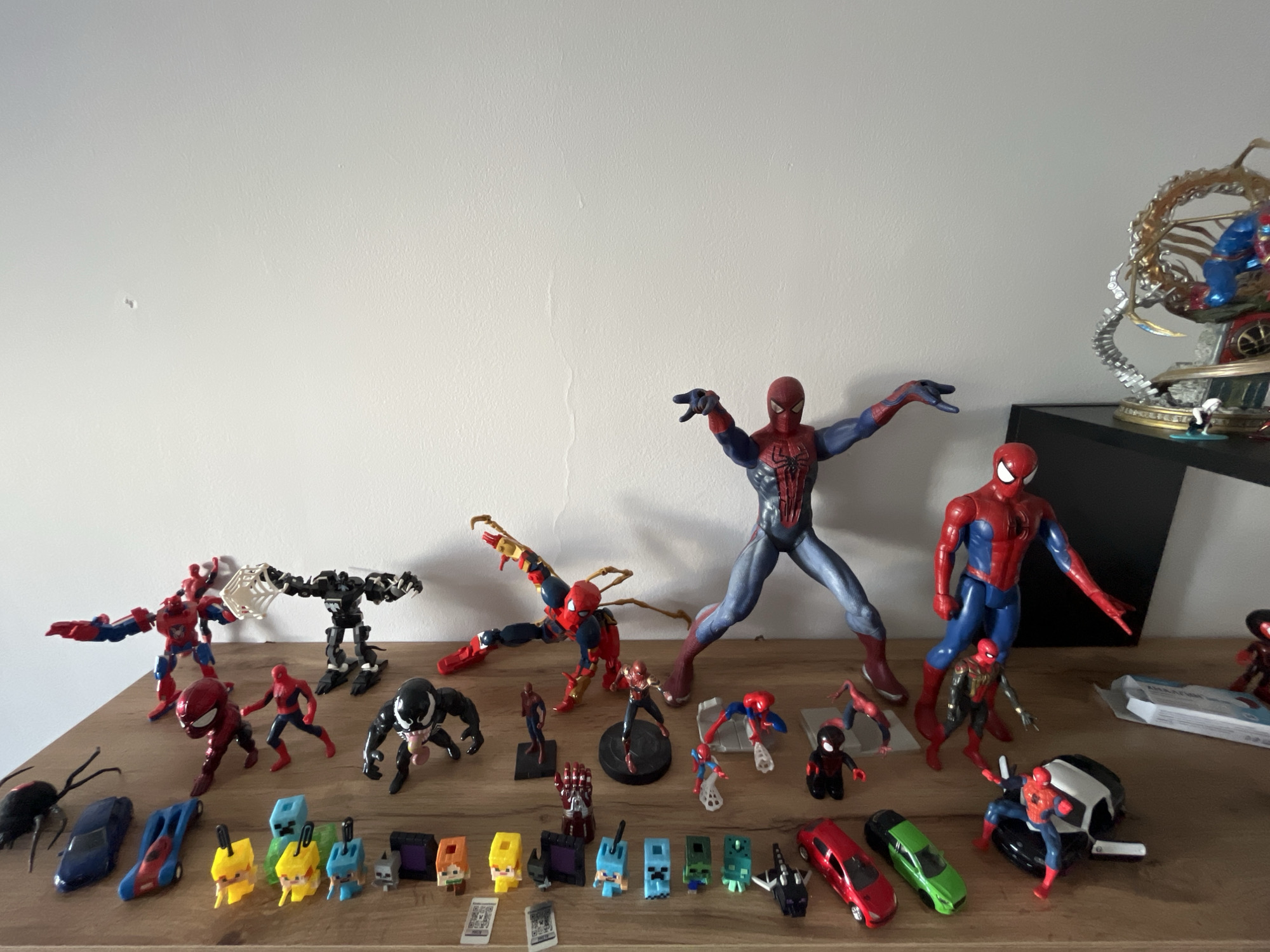 The image displays a collection of superhero action figures, primarily Spider-Man and Venom, arranged on a wooden shelf against a plain white wall. The figures vary in size and pose. Some are in dynamic action stances, while others stand more statically. In the background, there's a glimpse of a larger, more elaborate statue featuring multiple characters and metallic tentacles. The foreground of the shelf holds smaller items, including toy cars and what appear to be Minecraft-themed figures. The lighting suggests an indoor setting, likely a home or display area. There are no people visible in the image. The location of Shumen, Bulgaria is not indicated in the image.