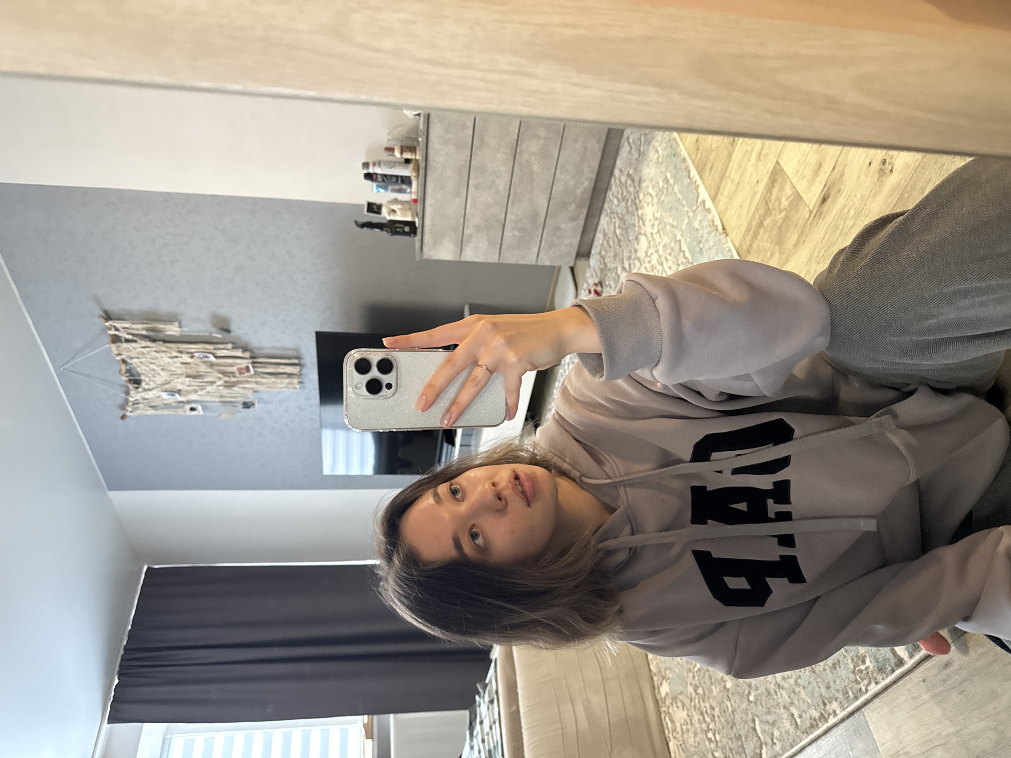 A woman is taking a selfie in a mirror. She is wearing a grey hoodie with the letters "GAP" on it. She is holding a silver iPhone in her right hand. Her hair is brown and she has green eyes. The reflection in the mirror shows a room with a grey wall, a white dresser, and a beige sofa. There are grey curtains in the background. It is not possible to determine from the image whether or not the location is in the city of Noviy, Russia.
