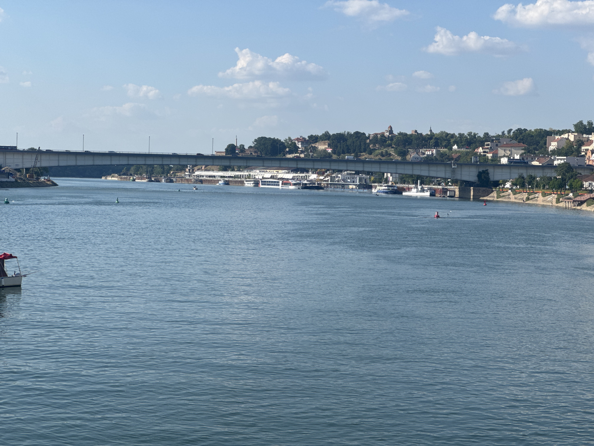 This media file captures a wide view of a river under a clear, blue sky with scattered white clouds, suggesting a pleasant daytime with fair weather. A large concrete bridge spans across the river, with traffic visible on its surface. Along the riverbanks, a mix of buildings, trees, and structures are visible, with some appearing to be part of a city or town. Several boats, including passenger vessels and smaller craft, are present on the water. One small boat with a red canopy is in the foreground on the left side of the image. The water itself is a calm, blue expanse with gentle ripples. The overall scene evokes a sense of a bustling urban waterway on a sunny day. The location is identified as Belgrade, Serbia, which aligns with the visual cues of the cityscape and riverine environment. There is no visible text in the image.
