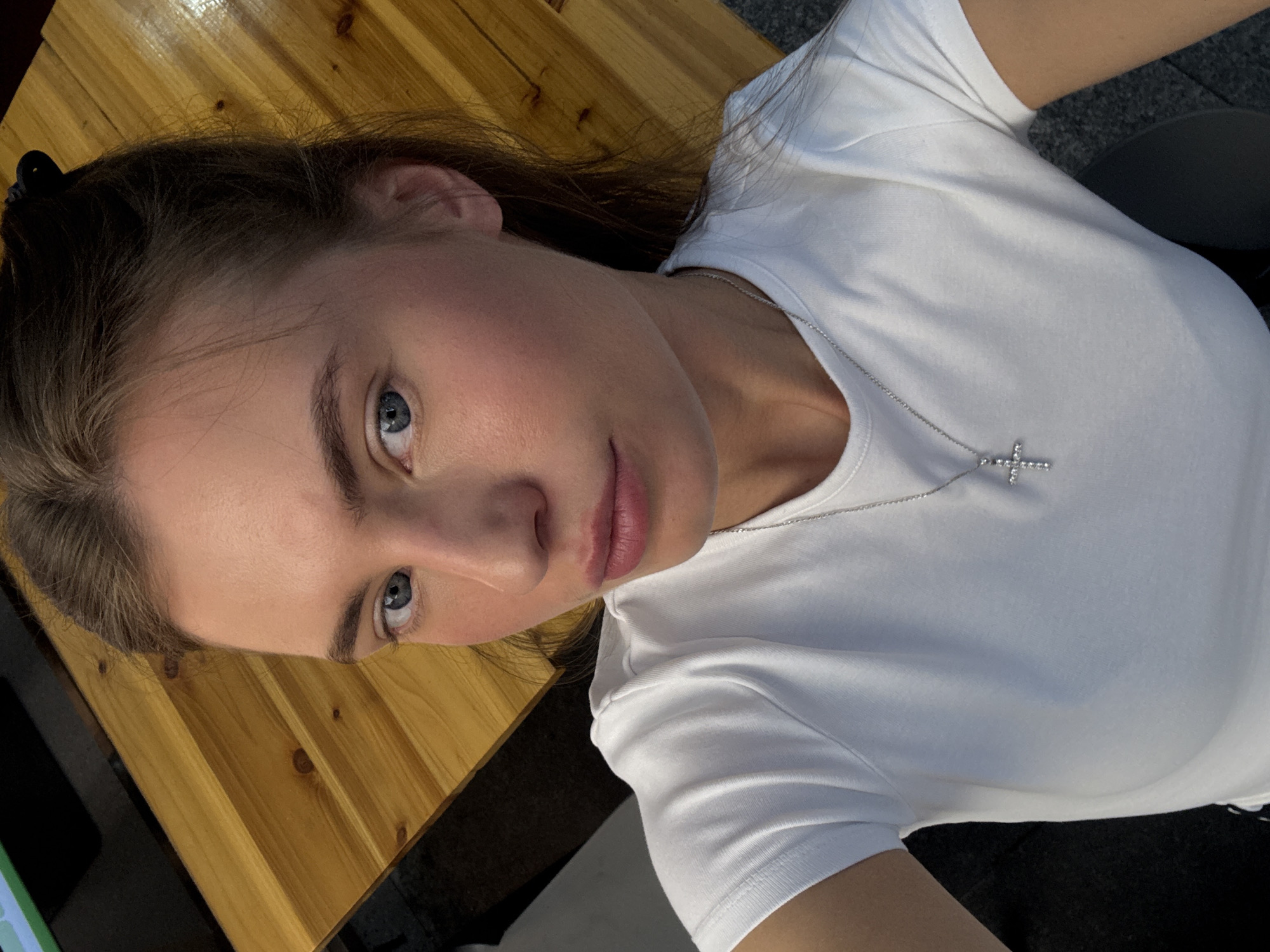 A young Caucasian woman with blue eyes and light brown hair is positioned in a close-up, upper-body shot, looking directly at the camera. She is wearing a white t-shirt and a silver necklace with a cross pendant. Her skin appears flushed, with noticeable blush on her cheeks. The background features a wooden surface, possibly a table, with distinct grain patterns. The image is taken indoors, under natural lighting that casts shadows. The overall impression is of a casual, everyday moment.