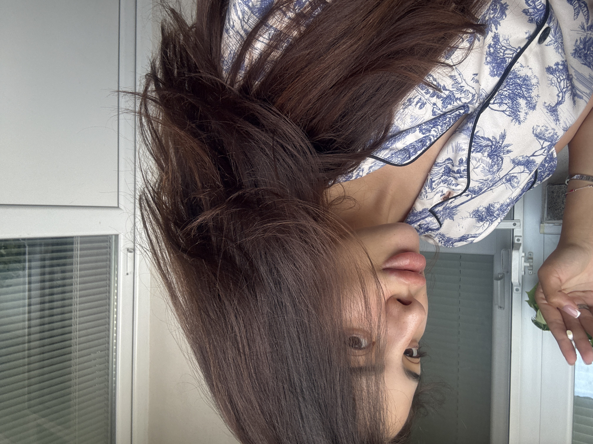 A woman with long, dark brown hair and light skin is shown in a close-up, profile view. She is wearing a white pajama top with a blue toile pattern and black trim. Her left hand, with manicured nails, is raised towards the left side of the frame, and she is wearing a ring on her left ring finger. The background is out of focus, showing what appears to be a window with blinds, suggesting an indoor setting. There are no discernible elements that place the image in Plovdiv, Bulgaria.