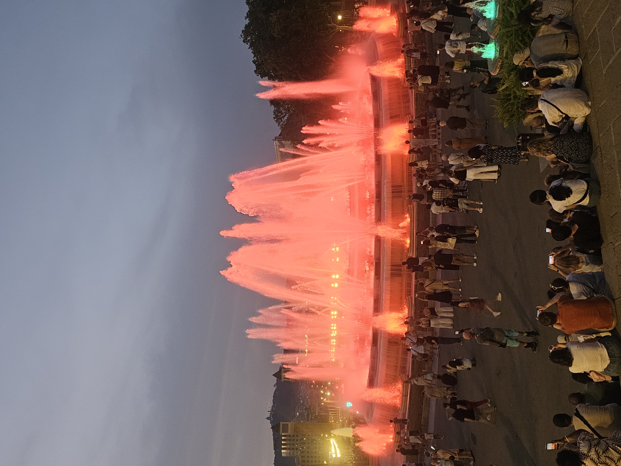 The image shows the Magic Fountain of Montjuïc in Barcelona, Spain at dusk. The large fountain is illuminated with red light, creating a vibrant spectacle of water jets. A crowd of people is gathered in front of the fountain, many of them holding up their phones to take pictures or videos. Some individuals are sitting on the steps leading up to the fountain, while others are standing. In the background, the cityscape of Barcelona is visible, with buildings and lights. Trees and streetlights are also present, contributing to the urban setting. The sky is a dusky blue, indicating the transition from day to night. The overall scene depicts a popular tourist attraction and a lively public gathering.