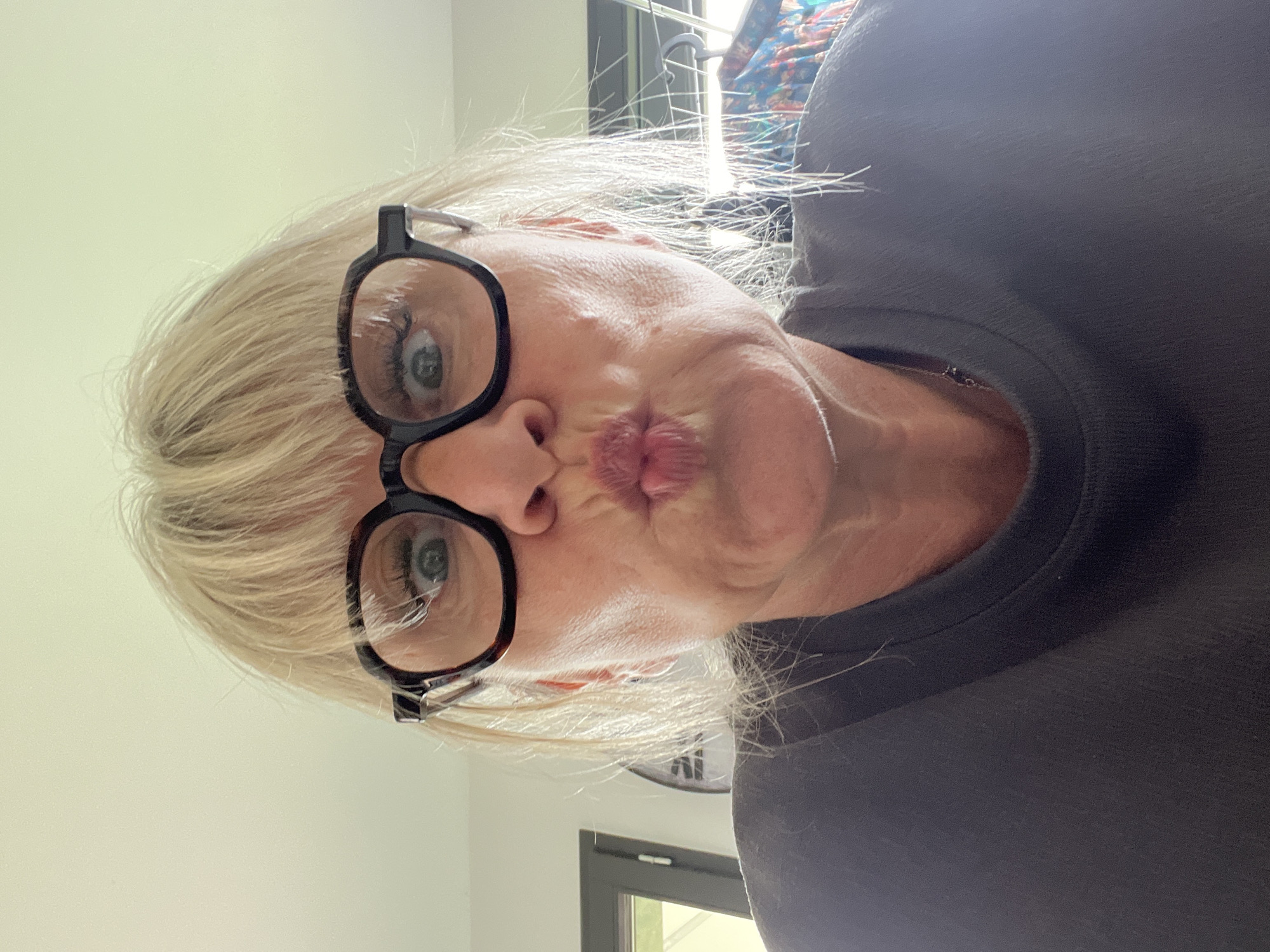 A woman with short blonde hair is centered in the image, wearing black-rimmed glasses and a dark-colored shirt. Her lips are pursed in a kissing gesture. The background includes a white wall on the left side and, behind her head, a clothes-drying rack. A dark-framed door with a visible green exterior is in the lower center of the image. A clock is present in the background as well. There are no other identifiable people, subjects, objects, actions, interactions, or environmental details to pinpoint the location in Chambœuf, France.