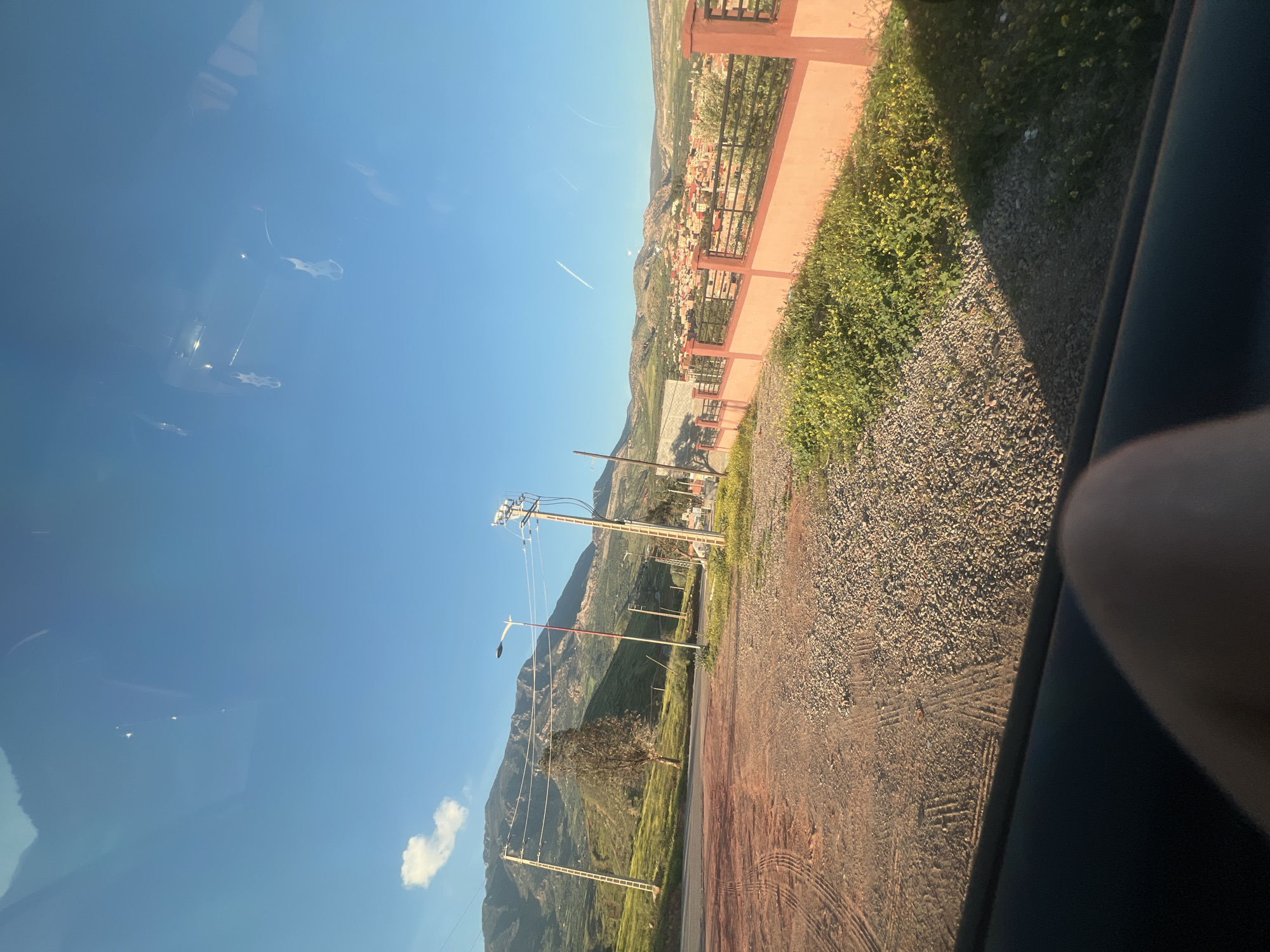 The image, taken from inside a vehicle, shows a road in Ouaouizert, Morocco. The road is unpaved and lined with gravel and vegetation. A pink painted wall with a metal fence runs alongside the road. Beyond the wall, a residential area of the town is visible, nestled in the hills. Power lines and poles are present. The sky is blue with a few clouds. Reflections are visible on the vehicle window. A portion of a person's hand or arm is visible inside the car.