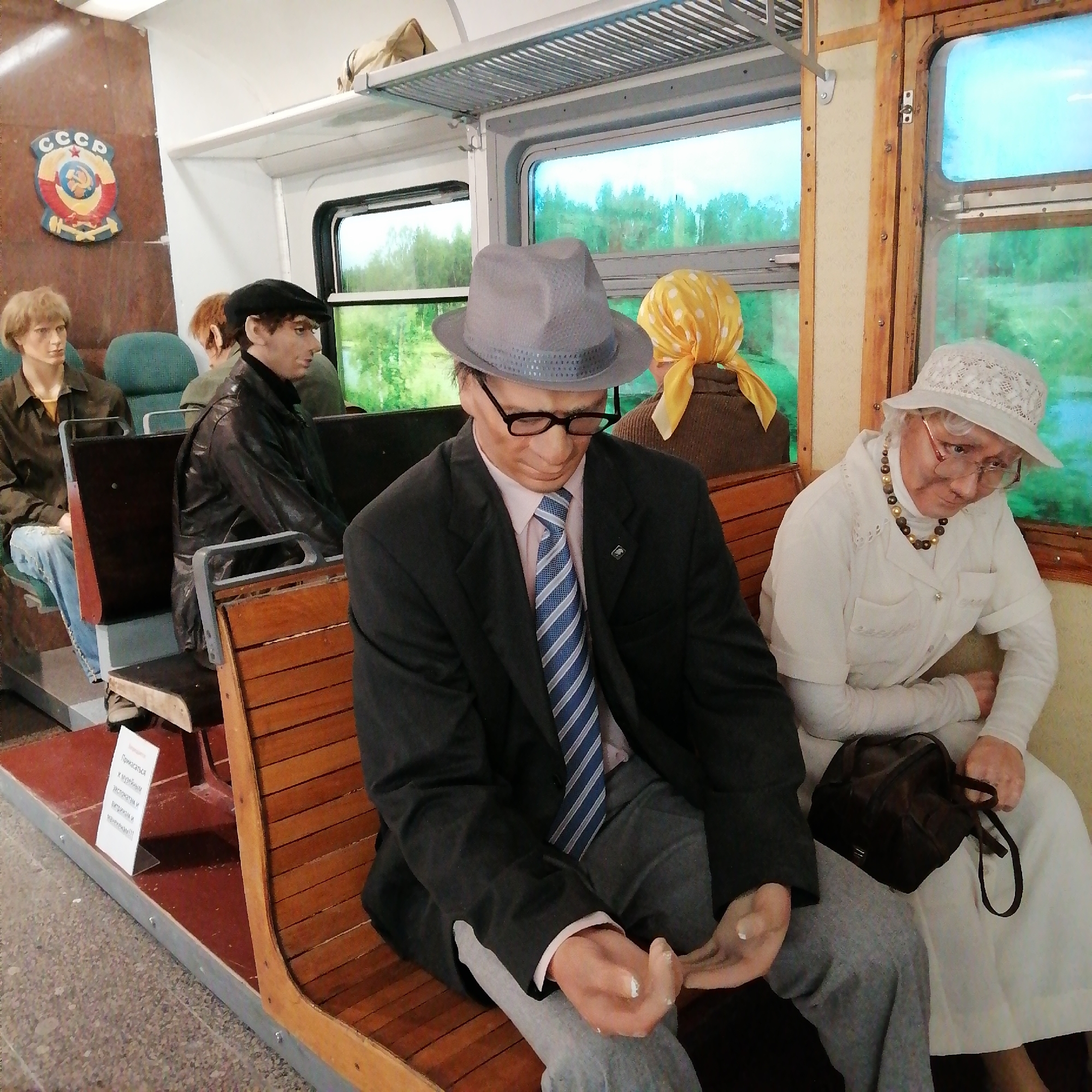 Inside a train car, mannequins of people sit on wooden benches. One male mannequin wears a gray fedora, glasses, a suit, and a striped tie. An older female mannequin wears a white dress, a white hat, and a necklace, with a brown purse on her lap. Other mannequins sit behind them, including one in a black leather jacket and a black hat. The train car has windows and a luggage rack above. A Soviet Union emblem is visible on the wall. A sign in Russian stands near the benches. The location is most likely a museum exhibit in Moscow, Russia, depicting a scene from the Soviet era.